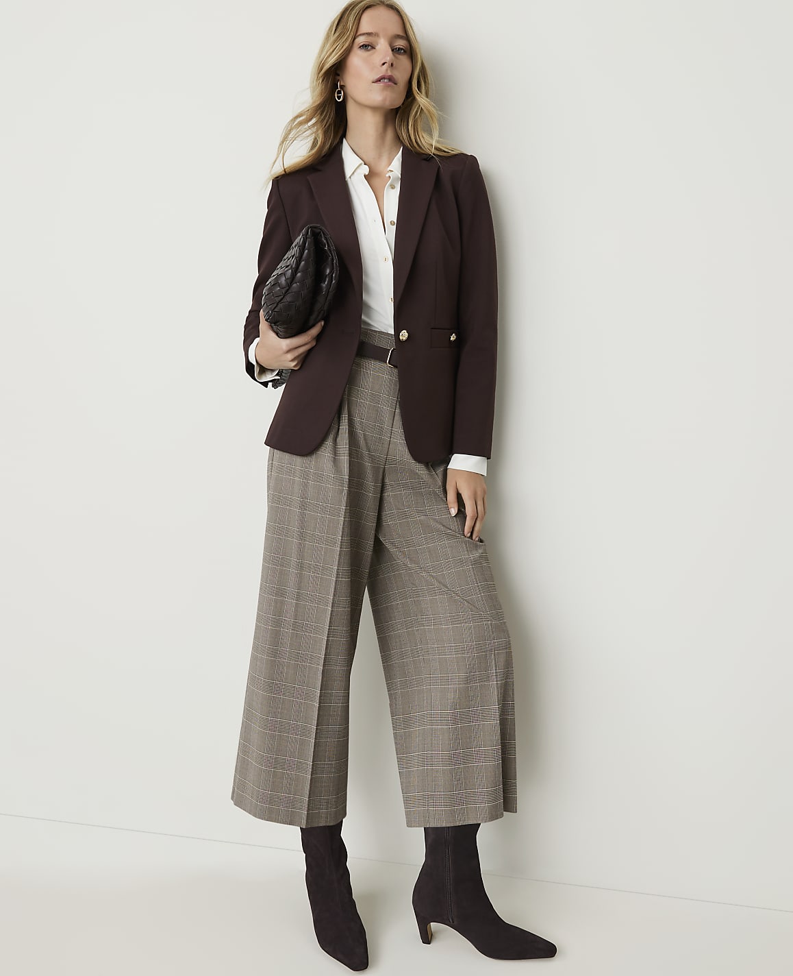 A woman in a brown blazer and plaid culottes stands against a plain white background, holding a black clutch.