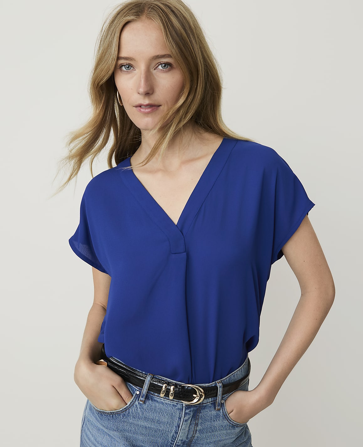A woman in a blue v-neck top and jeans stands against a plain white background.