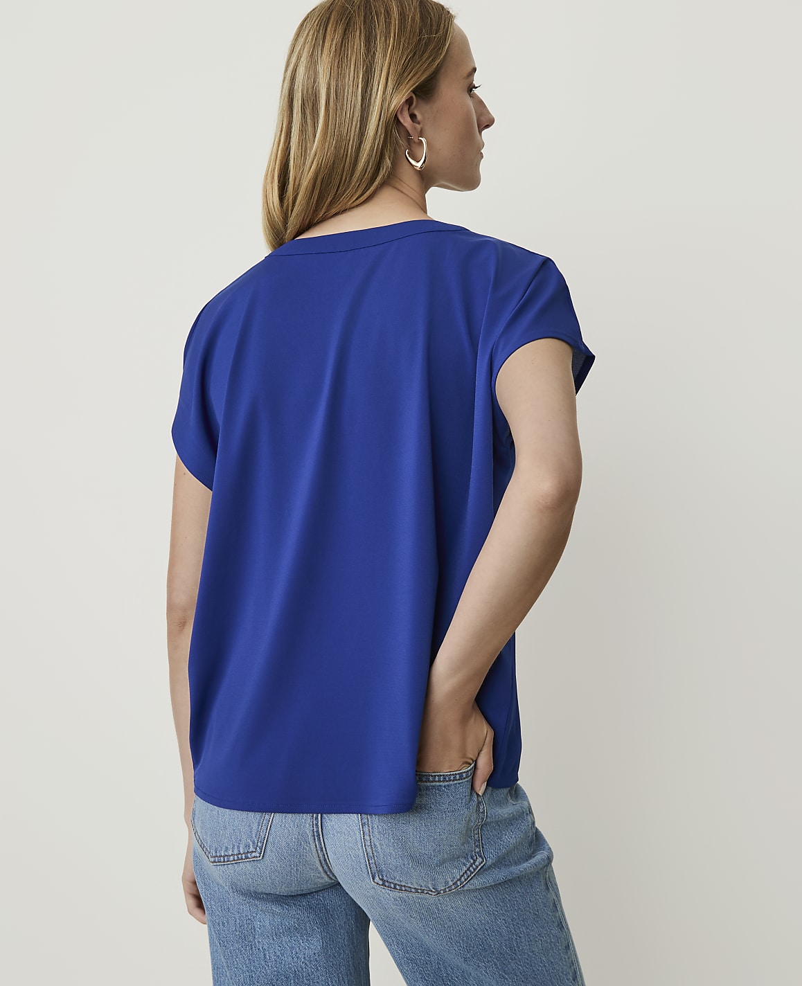 A woman in a blue t-shirt and jeans stands with her back to the camera against a plain white background.