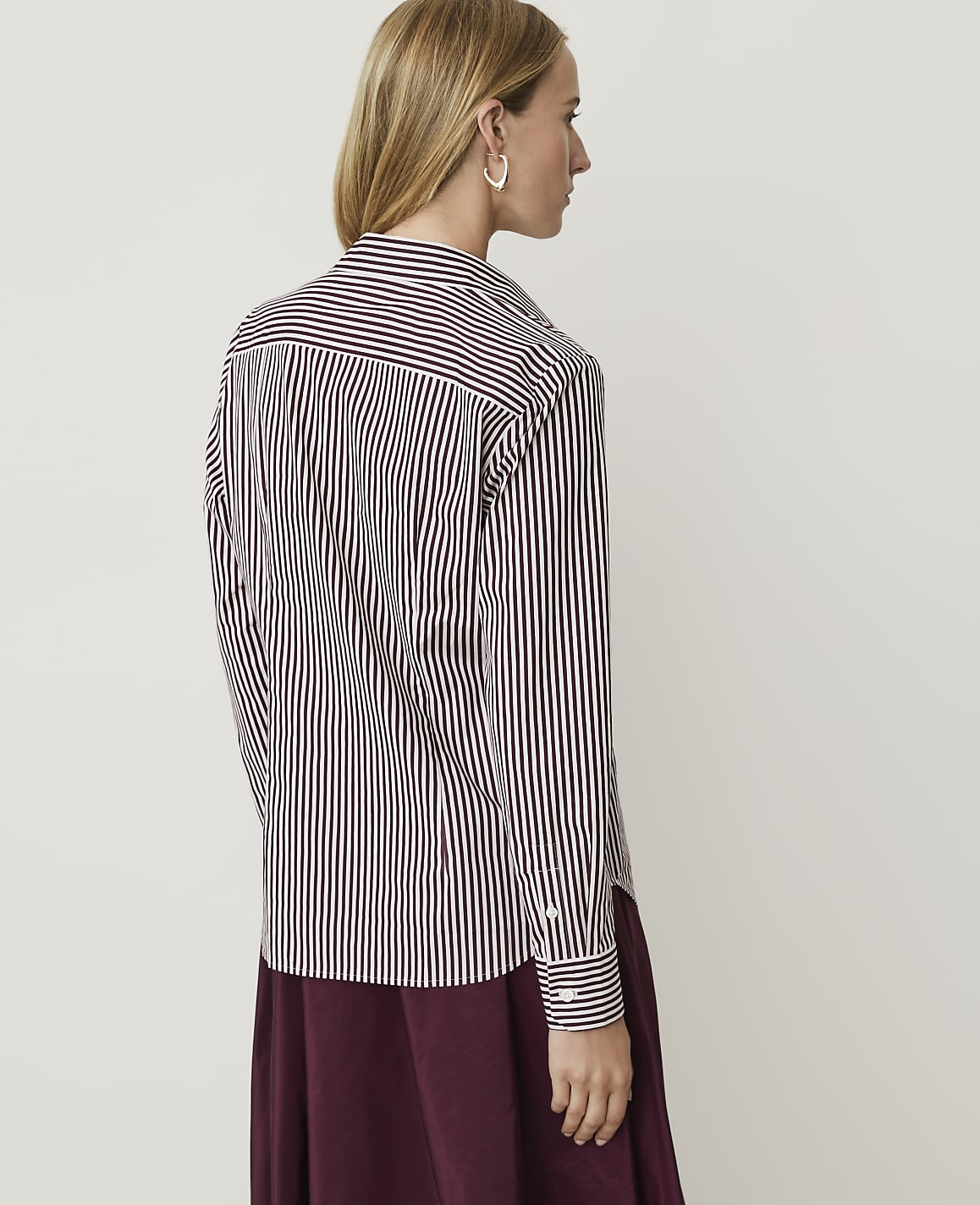 A woman in a striped shirt and maroon skirt stands against a plain white background.