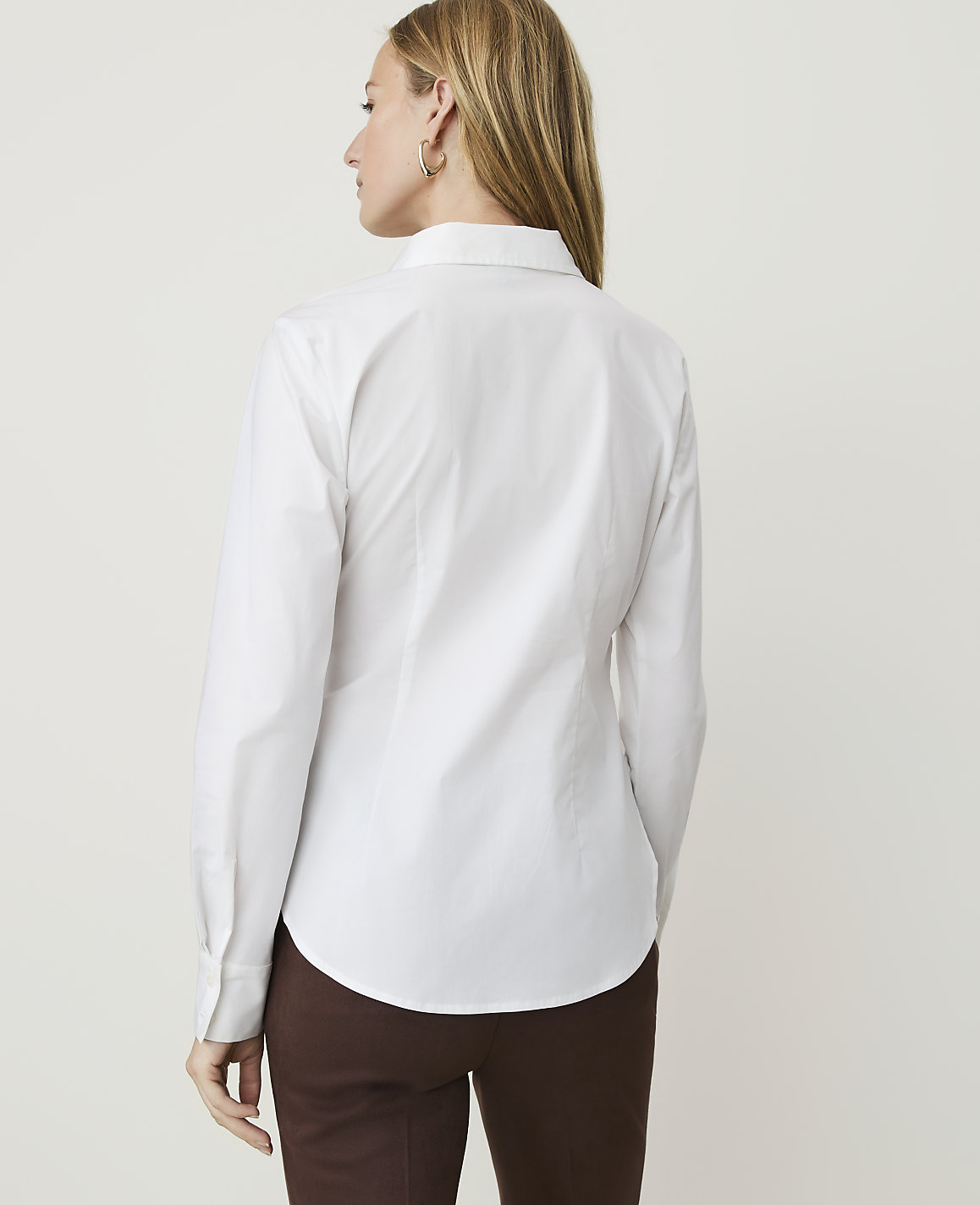 A woman in a white button-down shirt and brown pants stands against a plain white background.