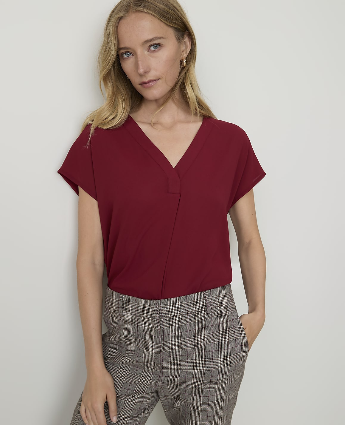A woman in a maroon v-neck top and plaid pants stands against a plain white wall.