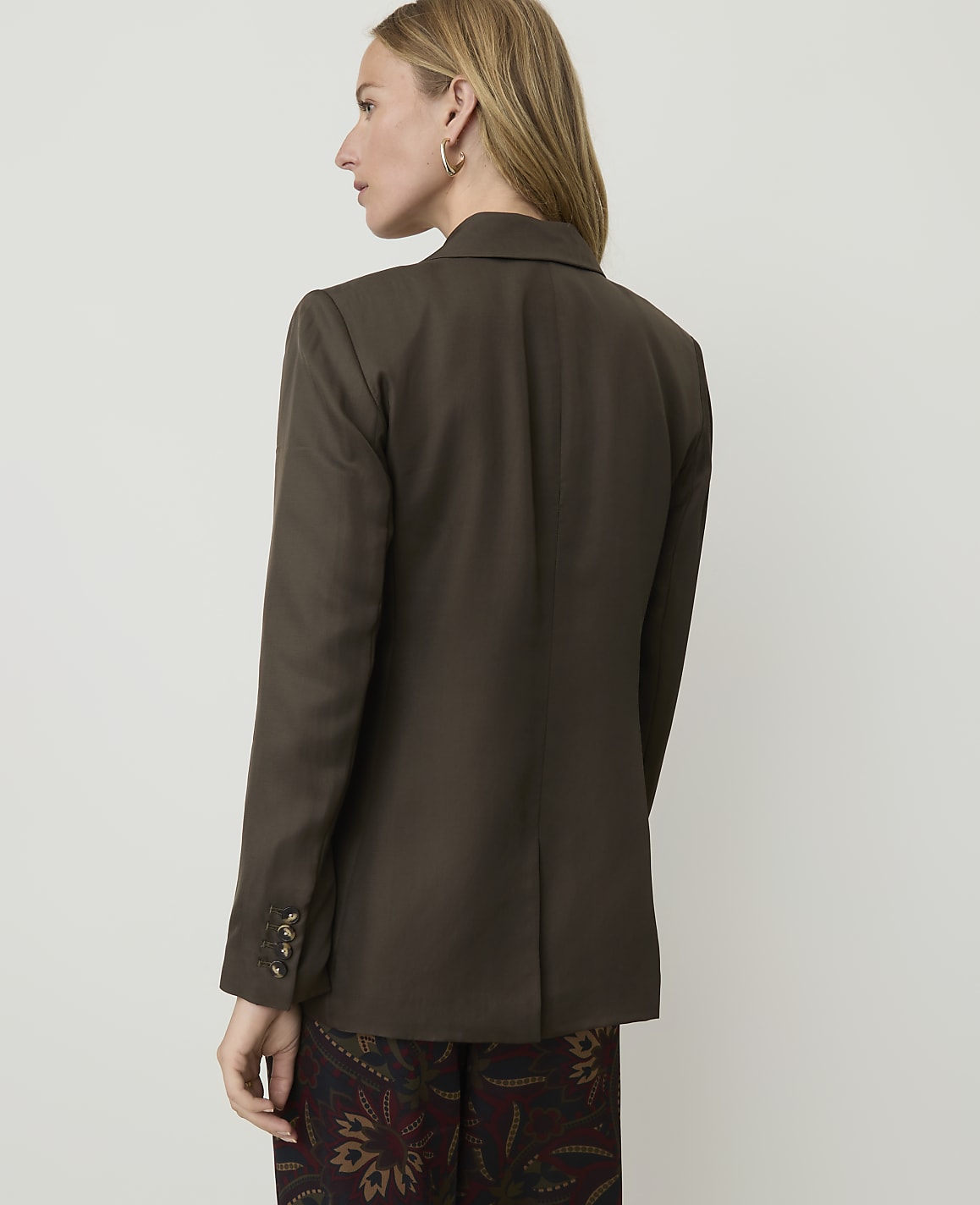 A woman in a brown blazer and patterned pants stands against a plain, light-colored background.