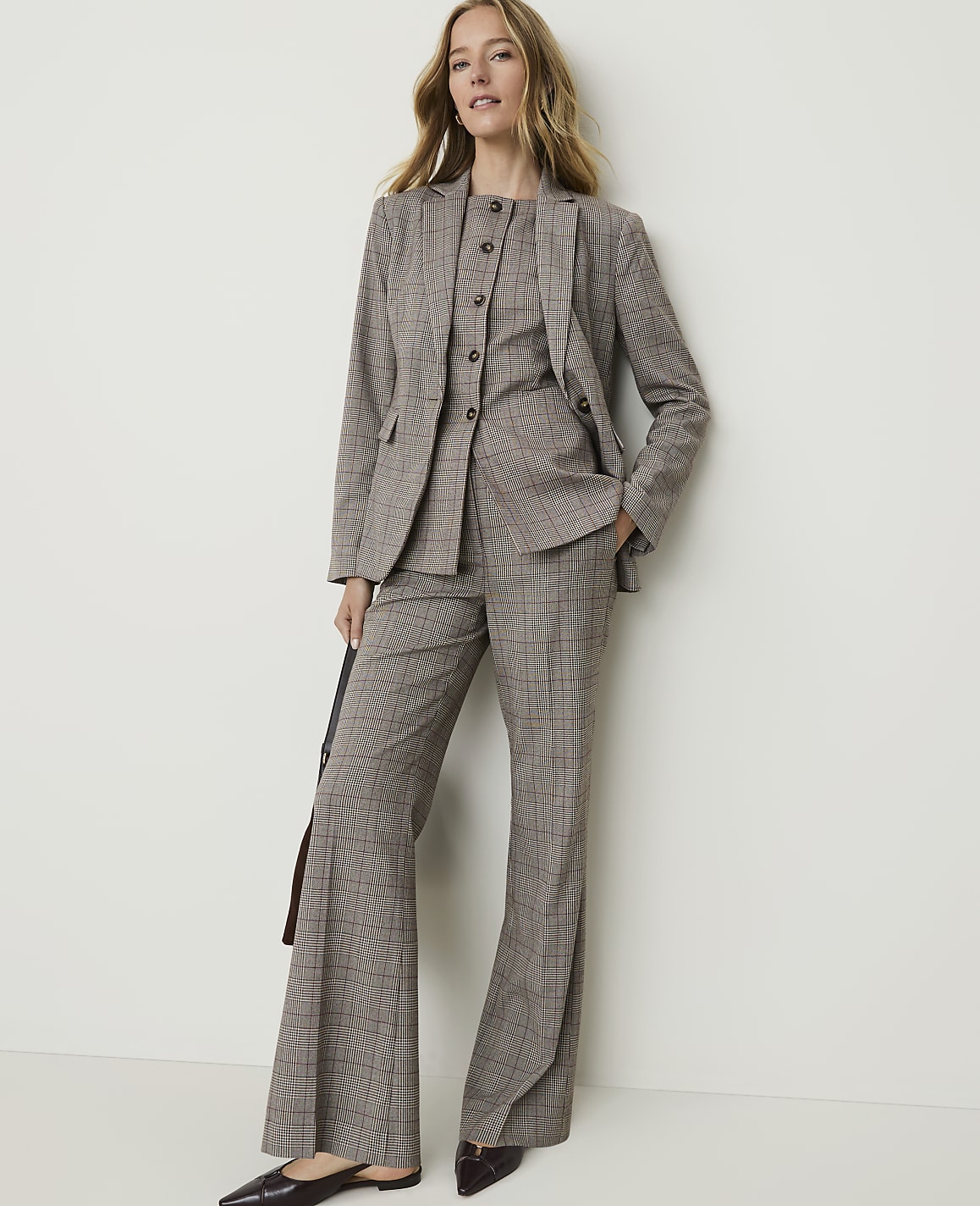 A woman in a brown plaid suit stands against a plain white wall, holding a dark bag.