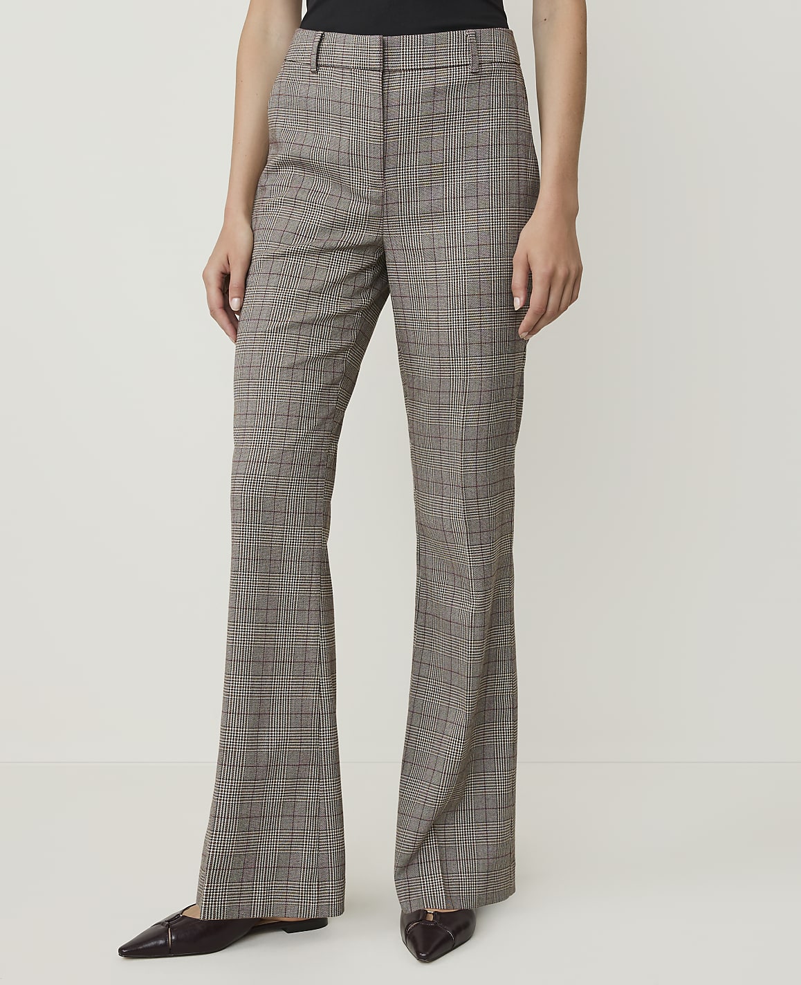 A person stands in the foreground wearing brown plaid flared trousers and dark pointed-toe shoes, against a plain white background.