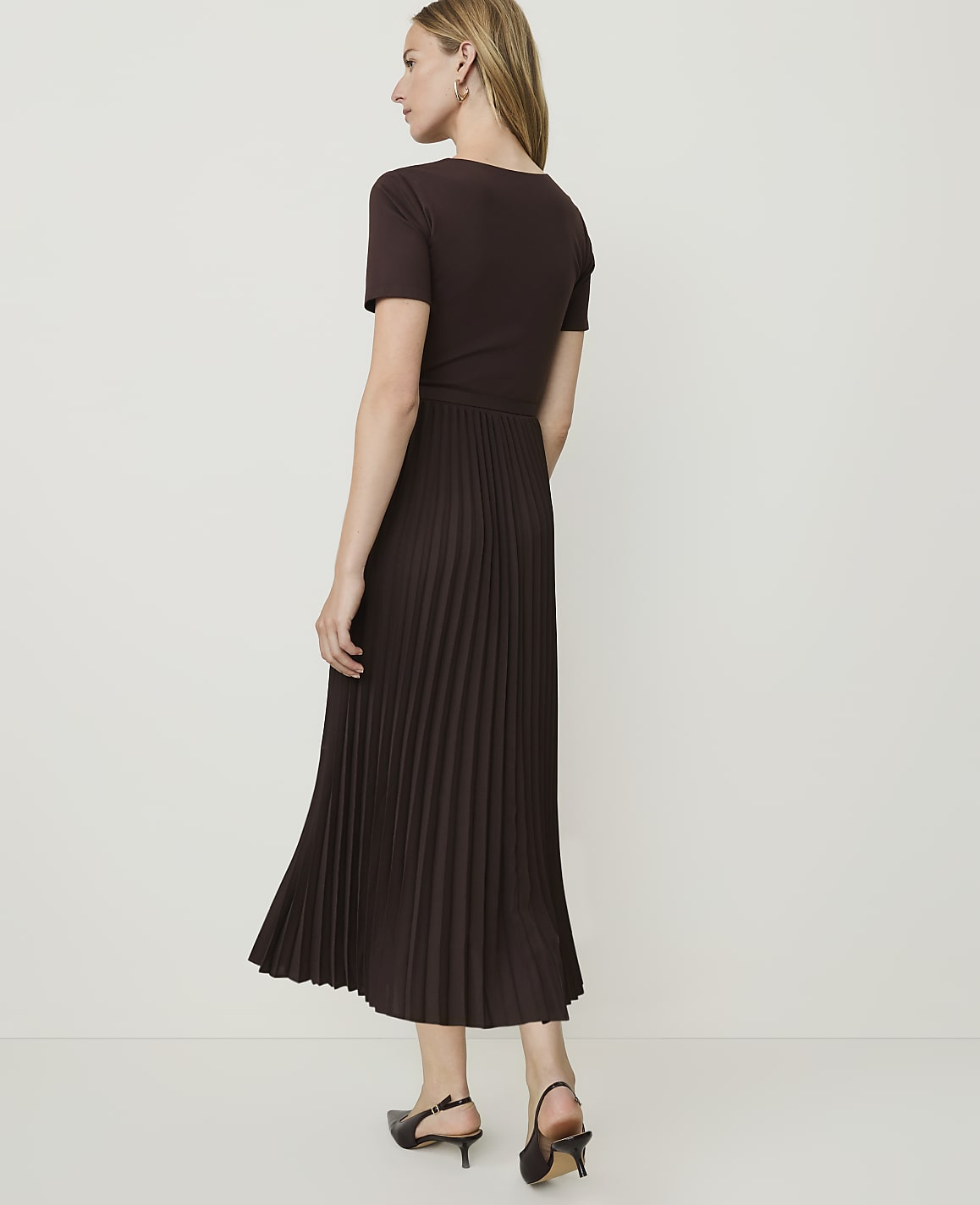 A woman in a brown t-shirt dress with a pleated skirt stands against a plain white background.