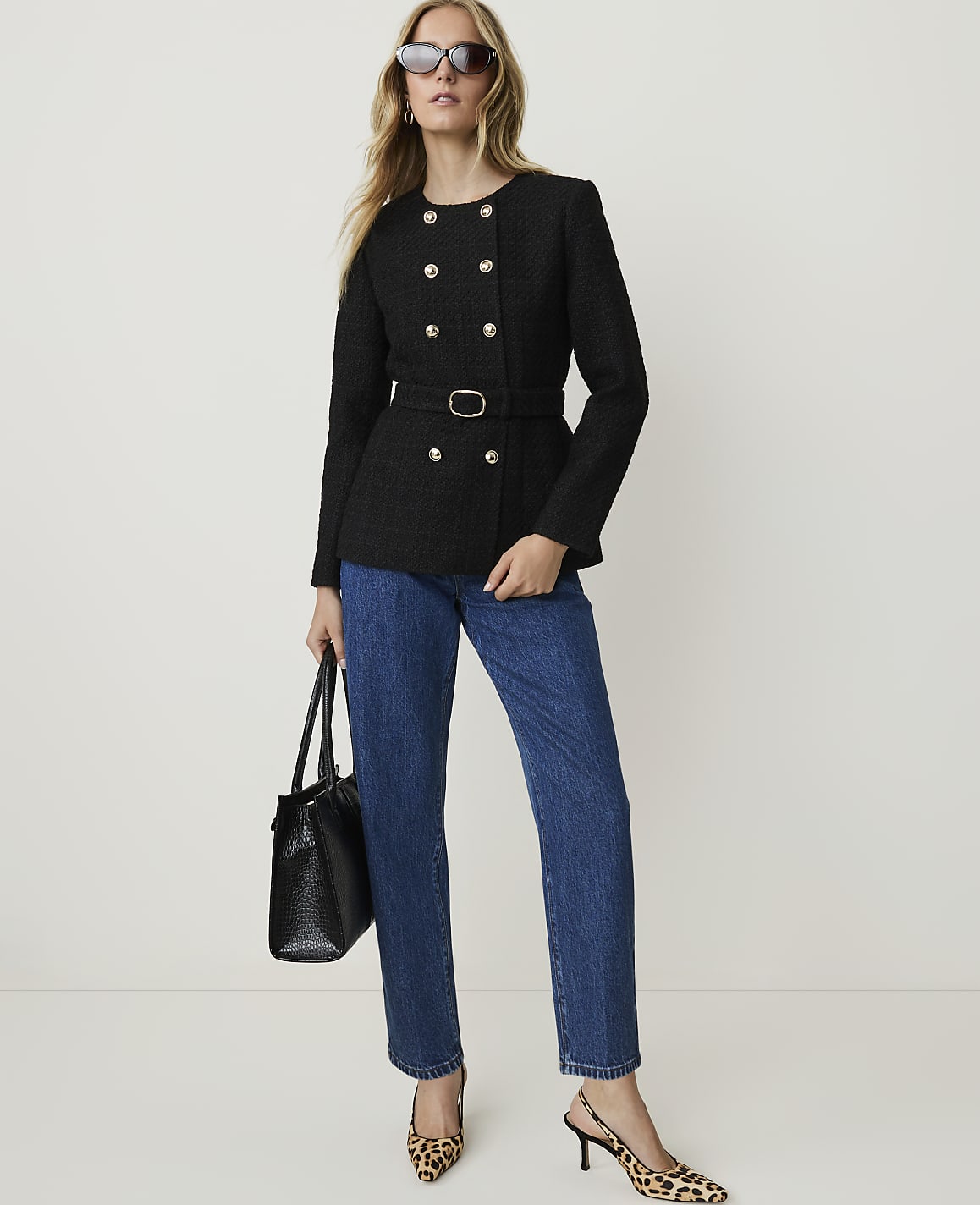 A woman in a black tweed jacket and blue jeans stands against a plain white background, holding a black handbag and wearing leopard print heels.