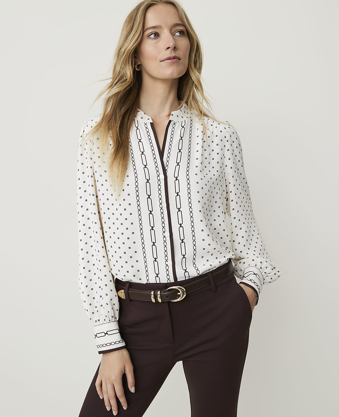 A woman in a patterned blouse and brown pants stands against a plain background.