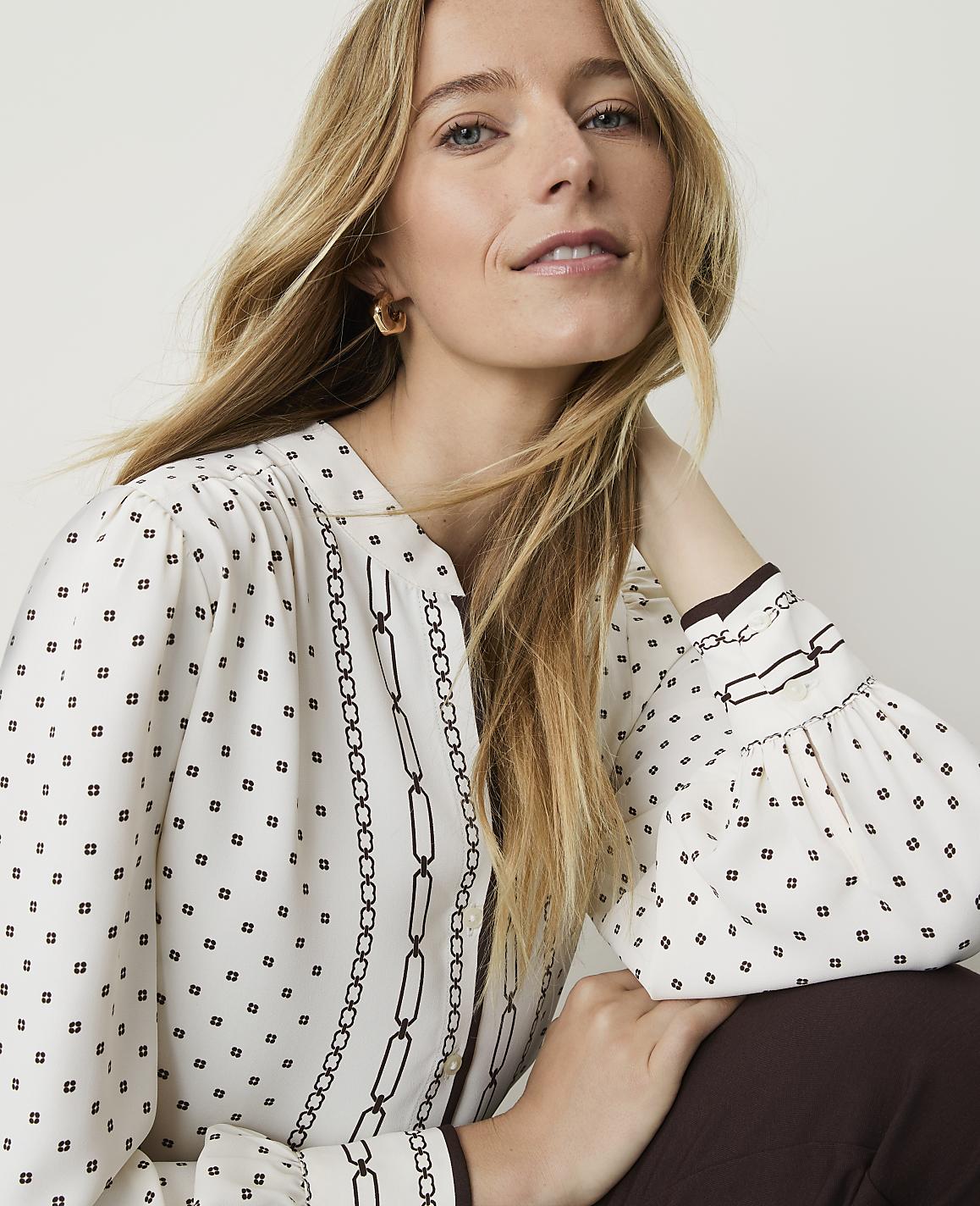 A blonde woman in a patterned blouse and brown pants poses against a plain background.