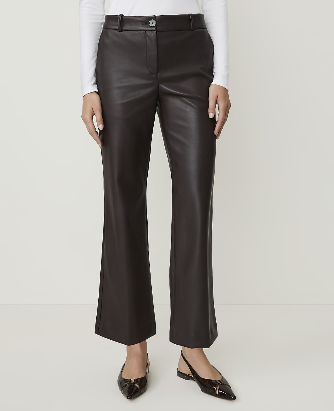 A person stands in dark brown faux leather pants and black pointed-toe flats against a plain white background.
