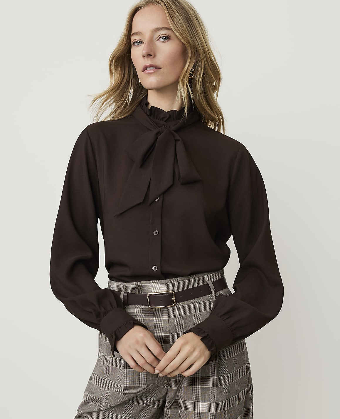 A woman in a brown pussy-bow blouse and plaid pants stands against a plain white background.