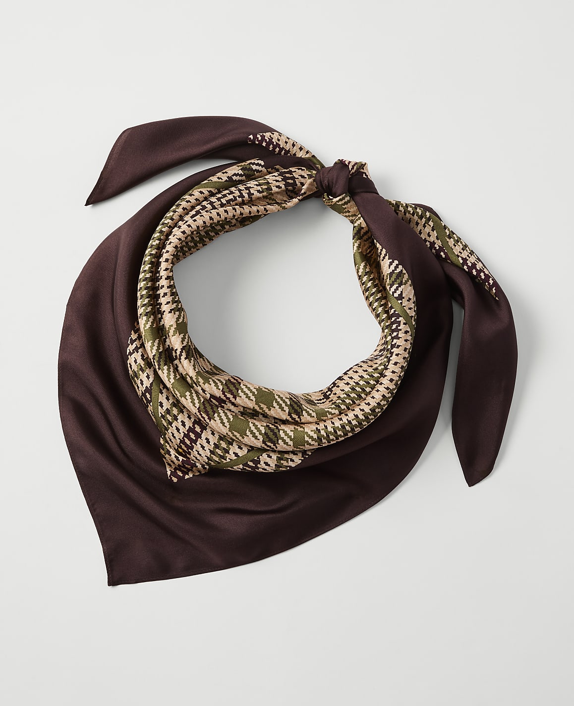 A brown silk scarf with a houndstooth pattern in green and beige is tied in a knot against a plain white background.