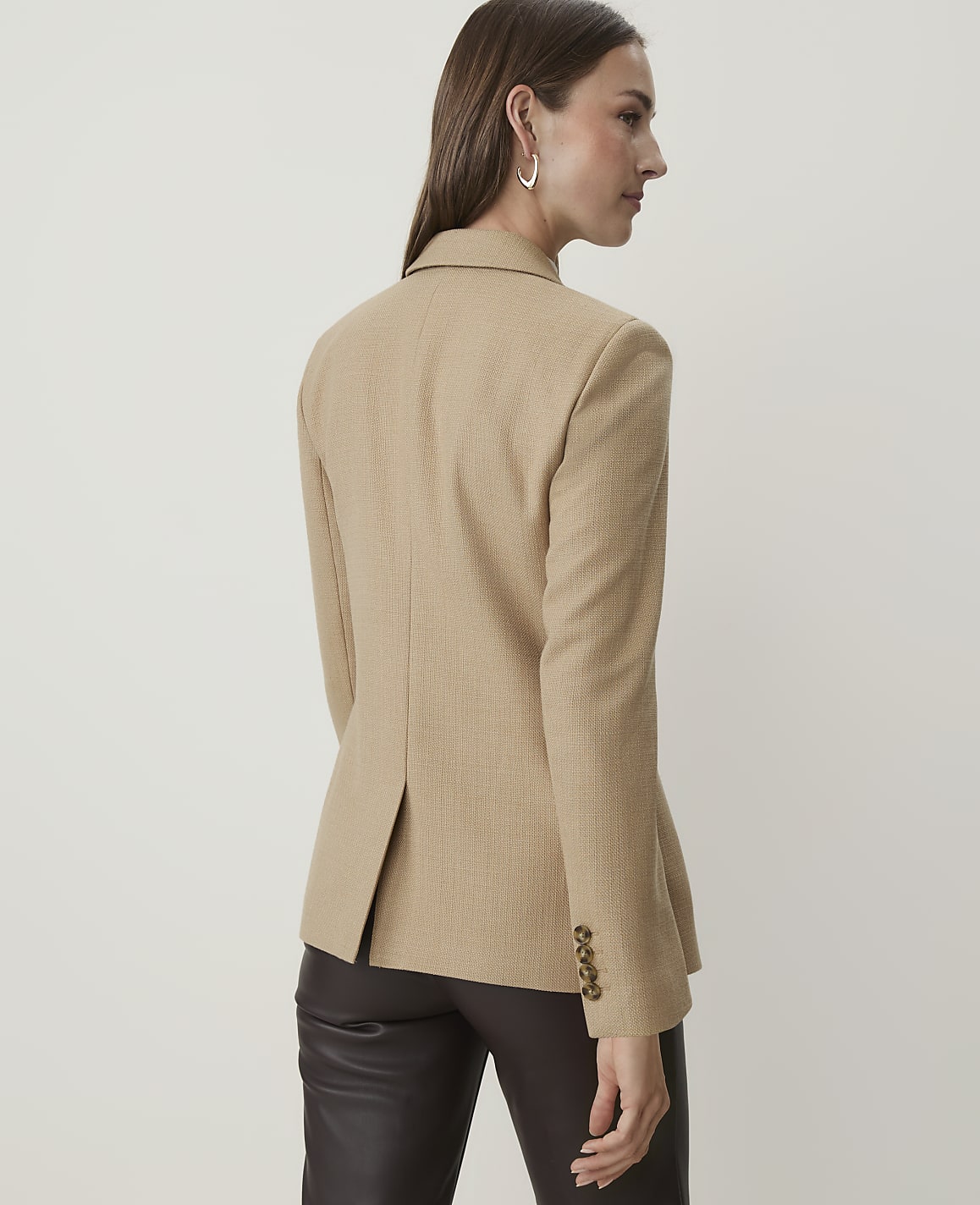 A woman in a tan blazer and dark pants stands against a plain, light-colored background.