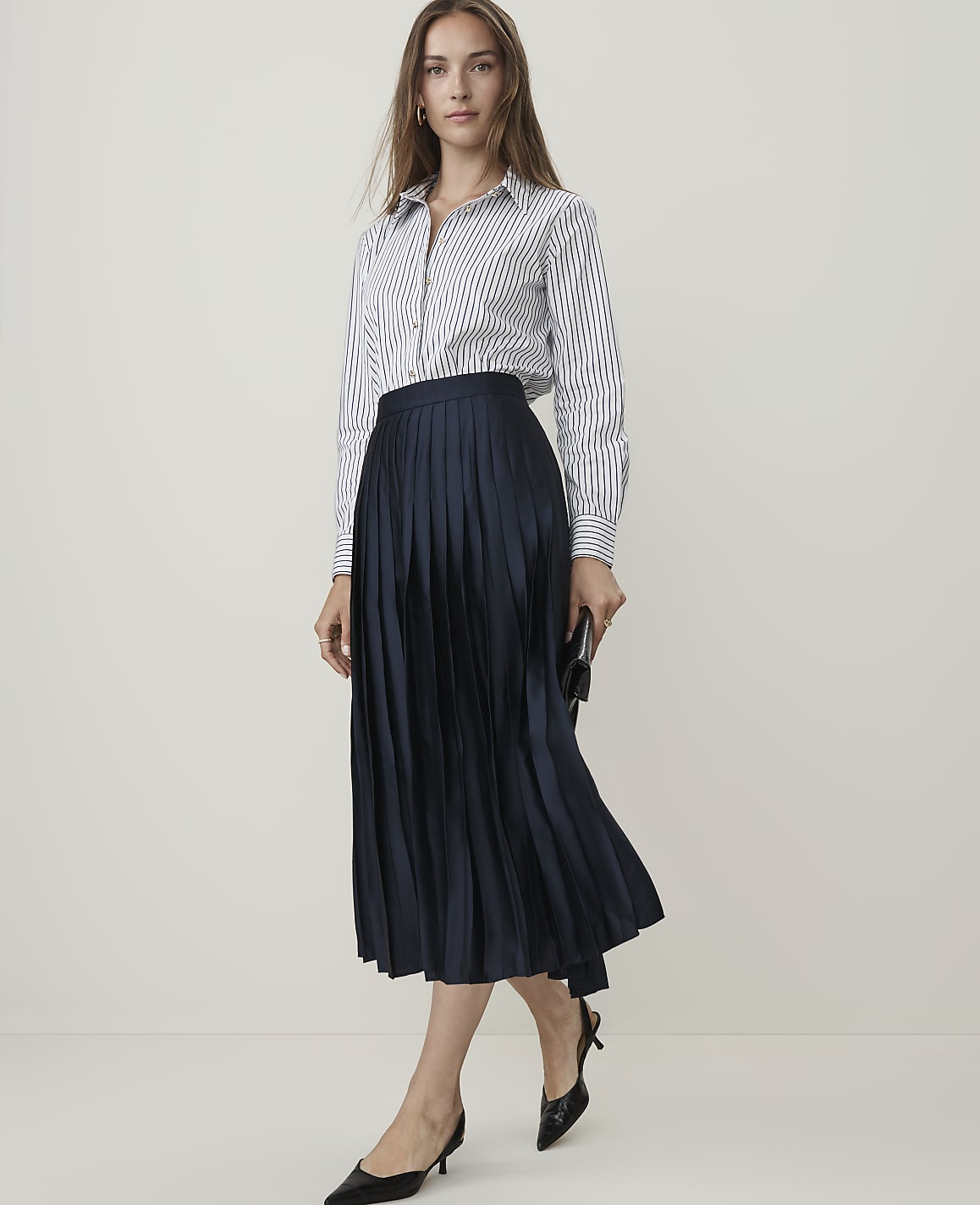 A woman in a striped shirt and navy pleated skirt stands against a plain, light background.