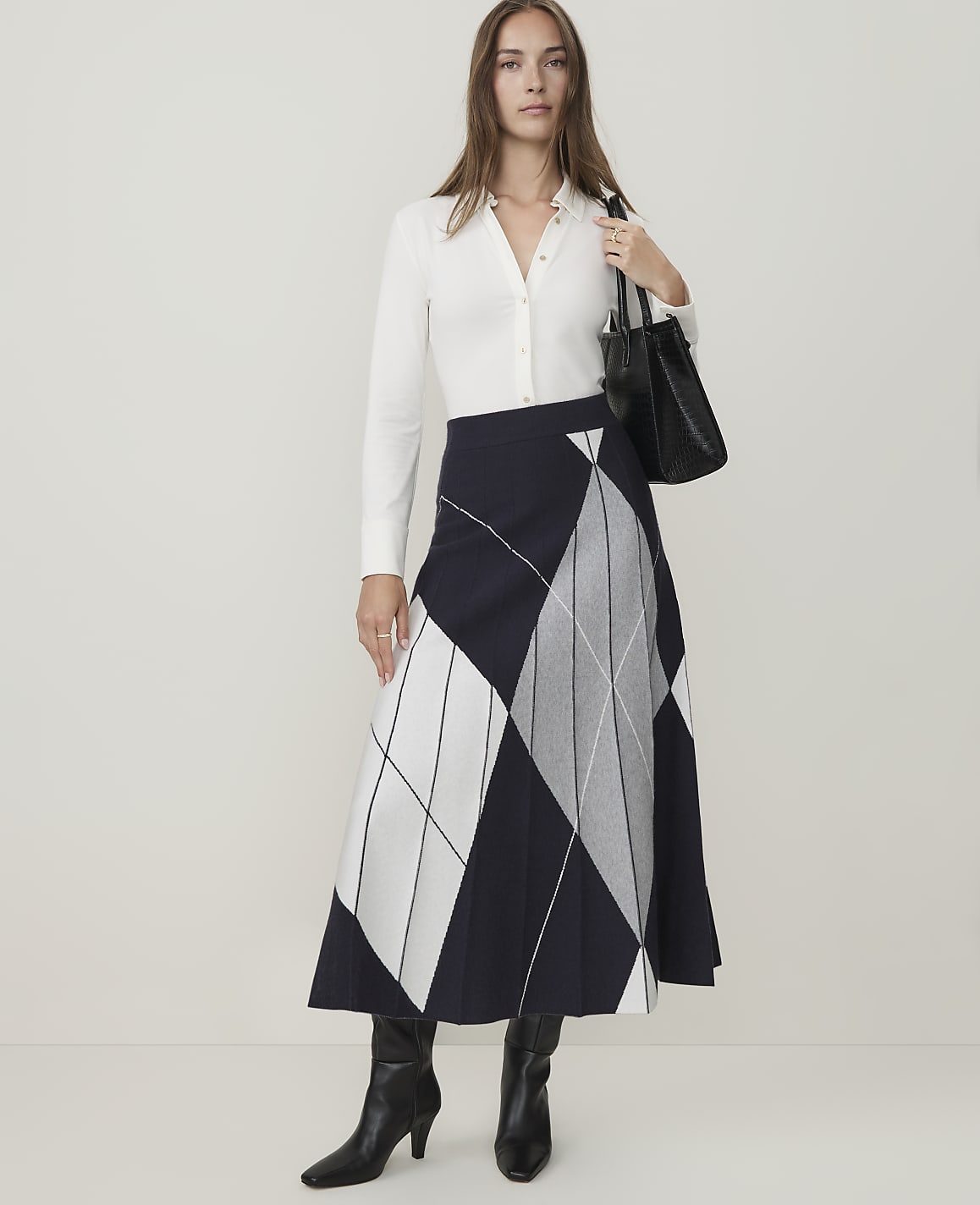 A woman in a white shirt, patterned skirt, and black boots stands against a plain white background, holding a black handbag.