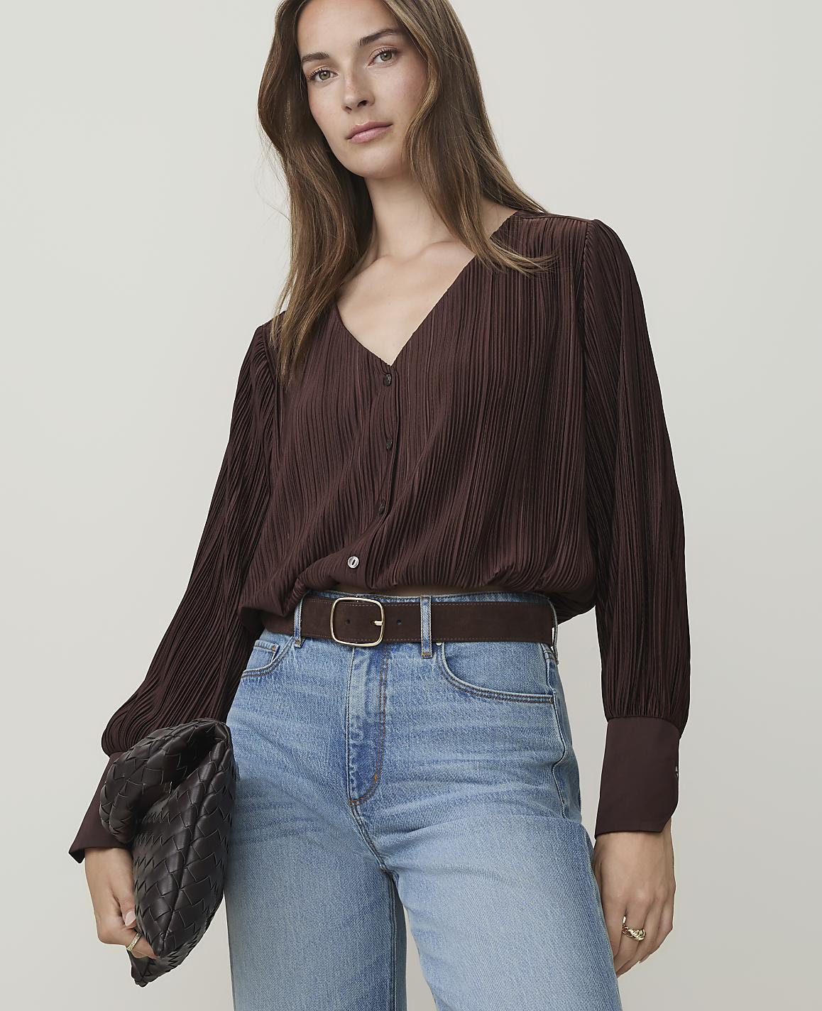 A woman in a brown pleated blouse and jeans holds a dark clutch in front of a plain background.