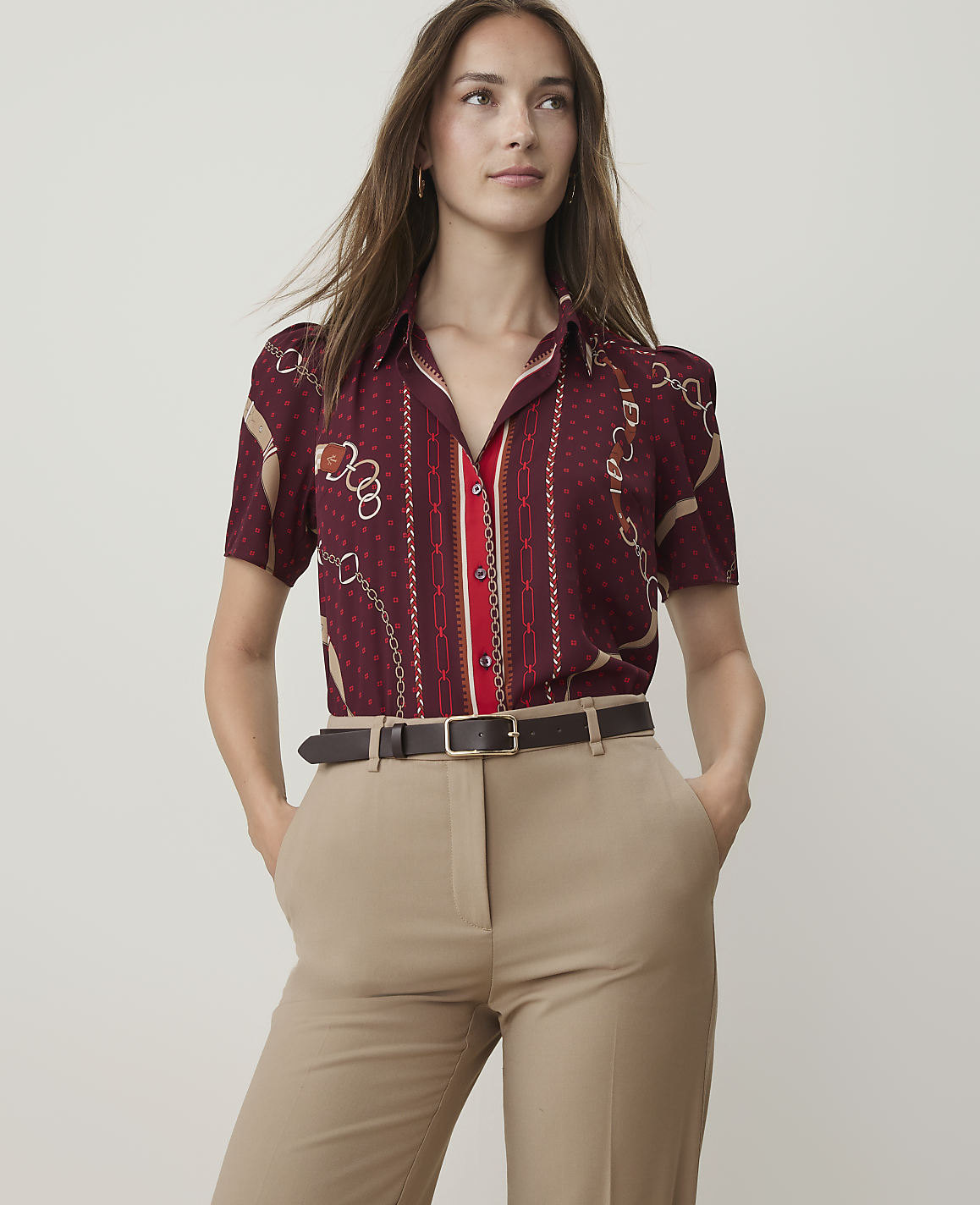 A woman in a patterned burgundy shirt and tan pants stands against a plain, light background.
