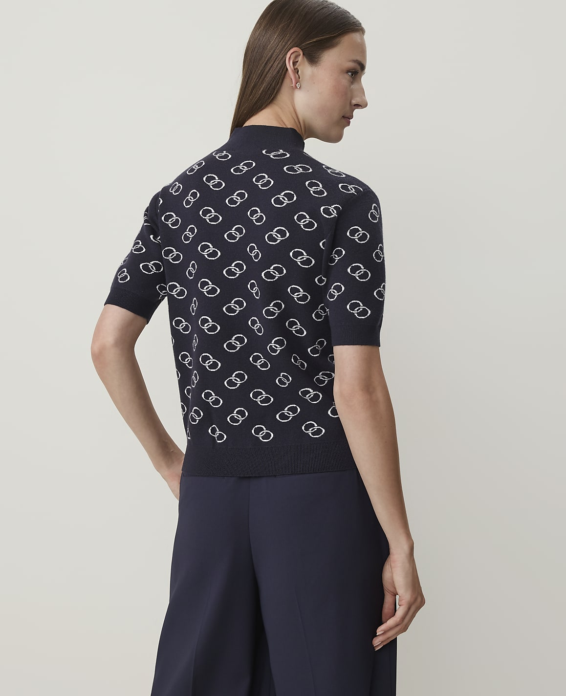 A woman in a dark blue, short-sleeved sweater with a white interlocking ring pattern stands against a plain, light gray background.