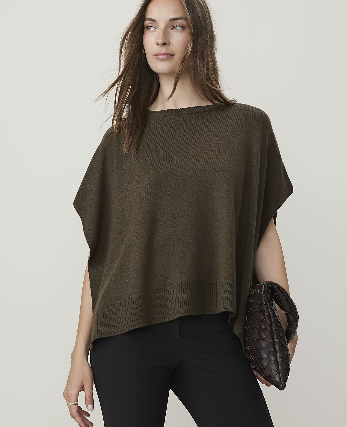 A woman in a dark olive poncho and black pants holds a woven clutch against a plain beige background.