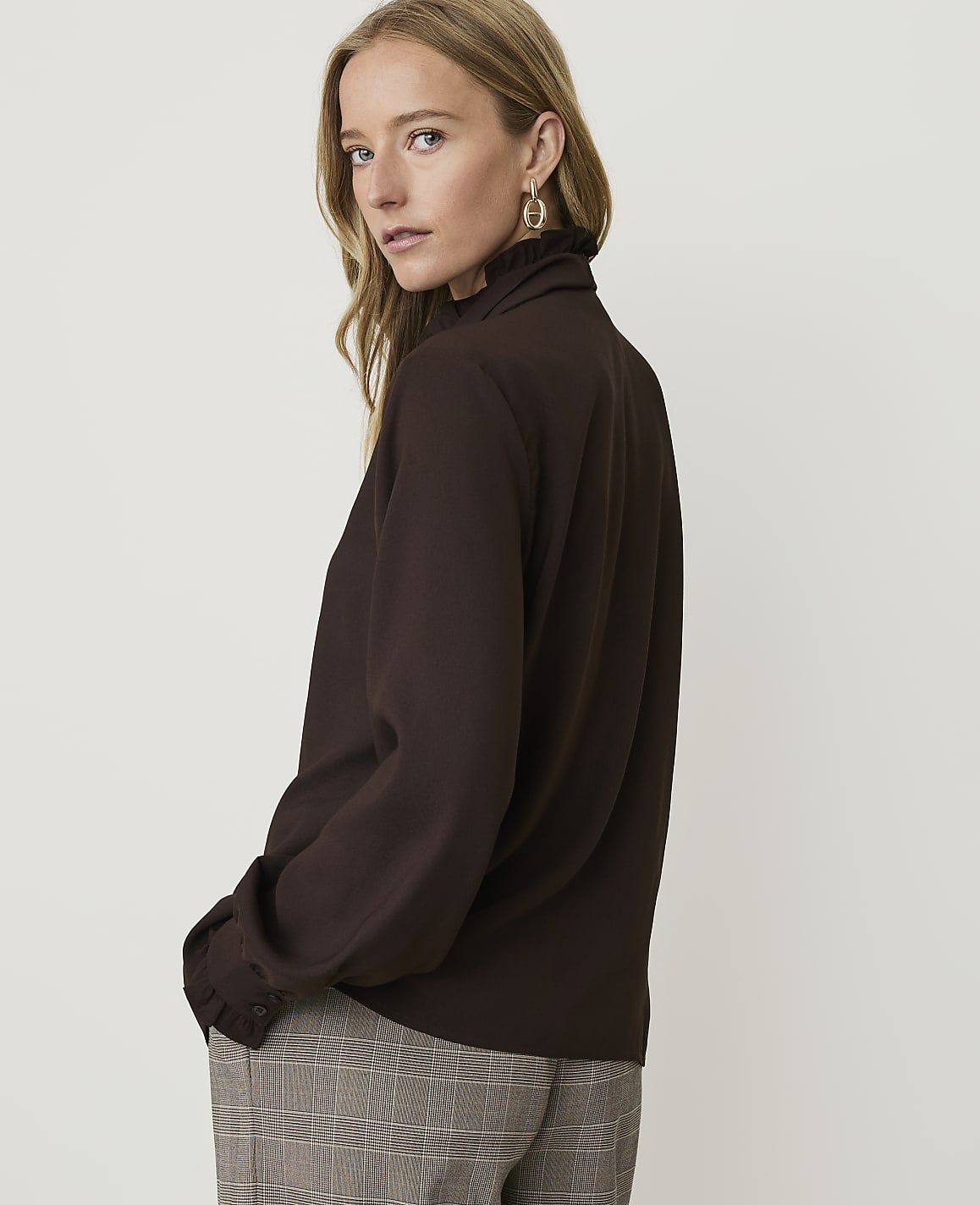 A woman in a brown ruffled-neck blouse and plaid pants stands against a plain, light brown background.