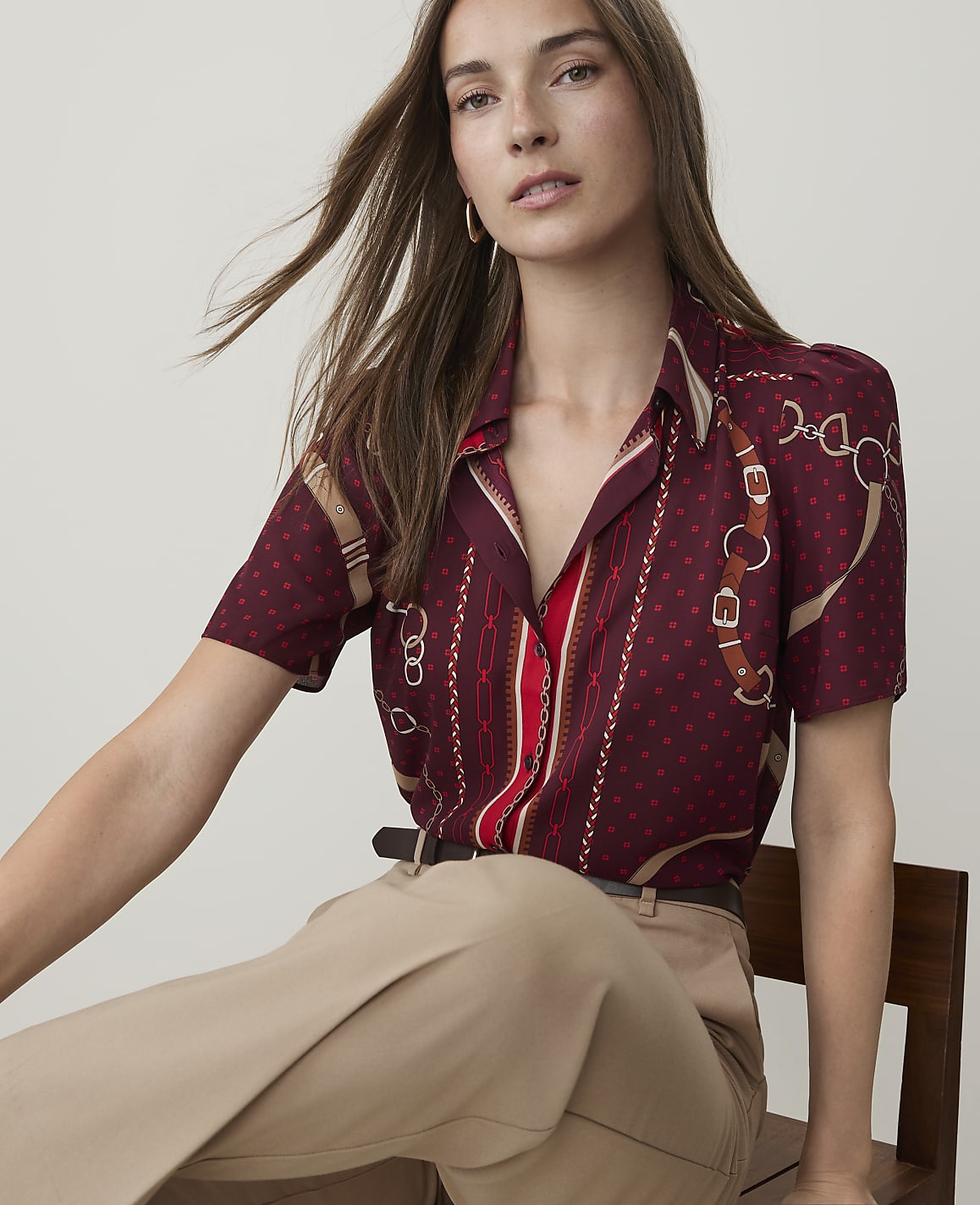 A woman in a patterned burgundy blouse and tan pants sits in front of a plain, light-colored background.