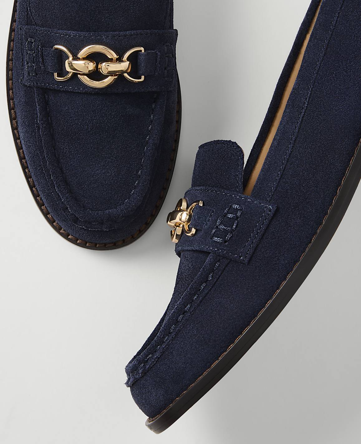 A pair of navy blue suede loafers with gold hardware rests on a plain white background.