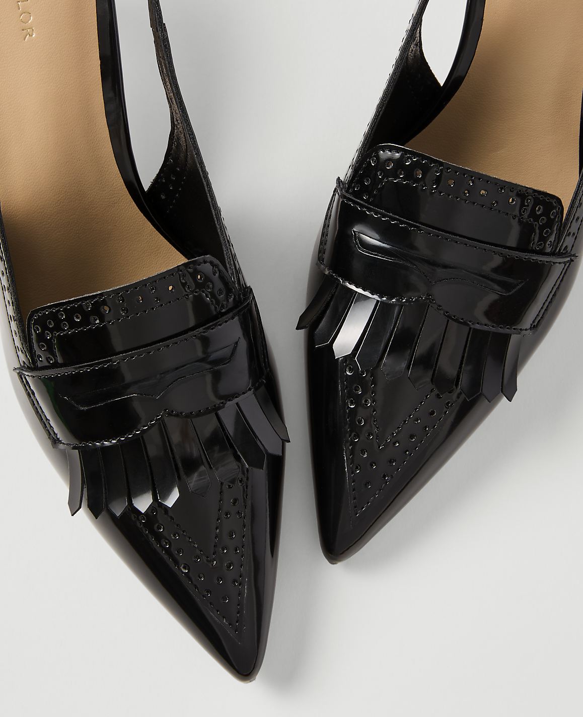 A pair of glossy black patent leather slingback heels with fringe detailing and brogue accents are presented against a plain white background.