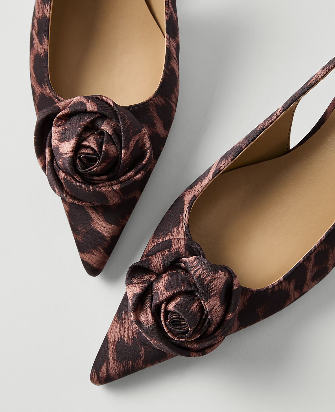 A pair of pointed-toe slingback heels with a leopard print and fabric rose embellishments are displayed against a plain white background.
