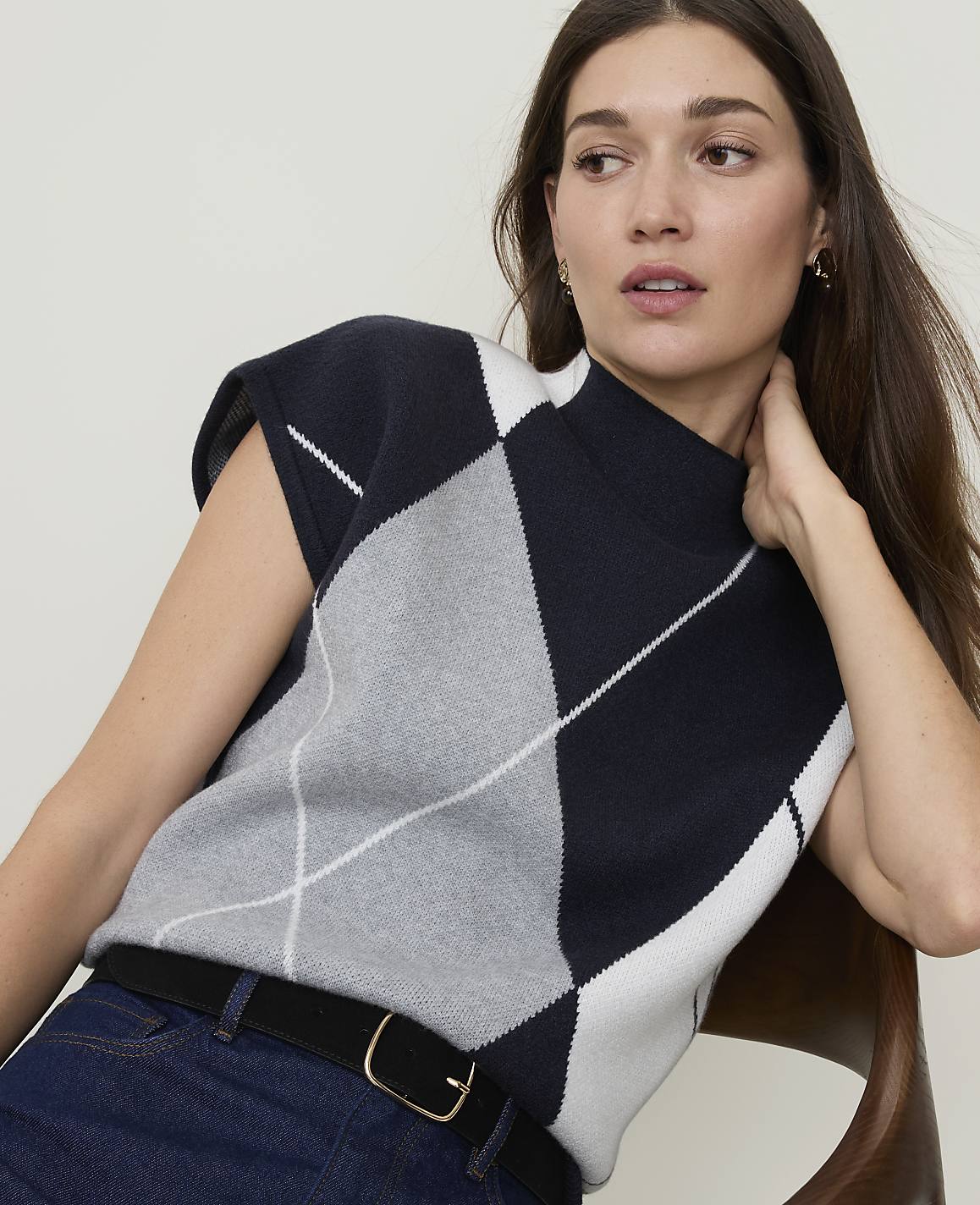 A woman in a sleeveless argyle sweater and dark jeans sits in front of a blurred background.