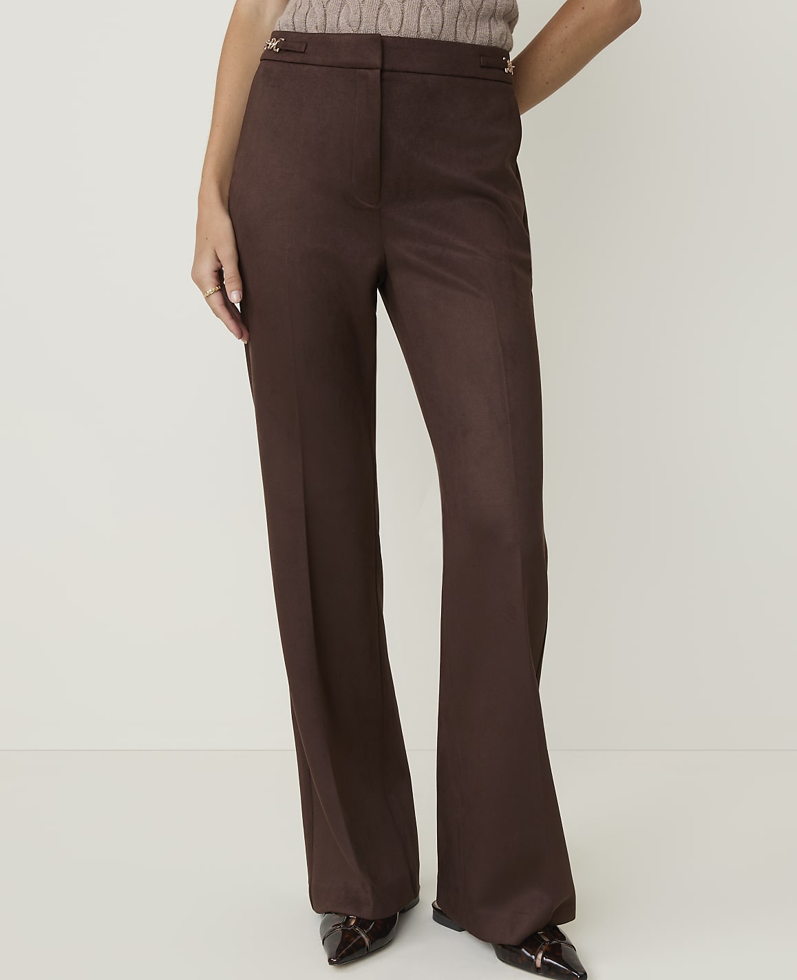 A person stands in brown flared trousers and pointed shoes against a plain white background.