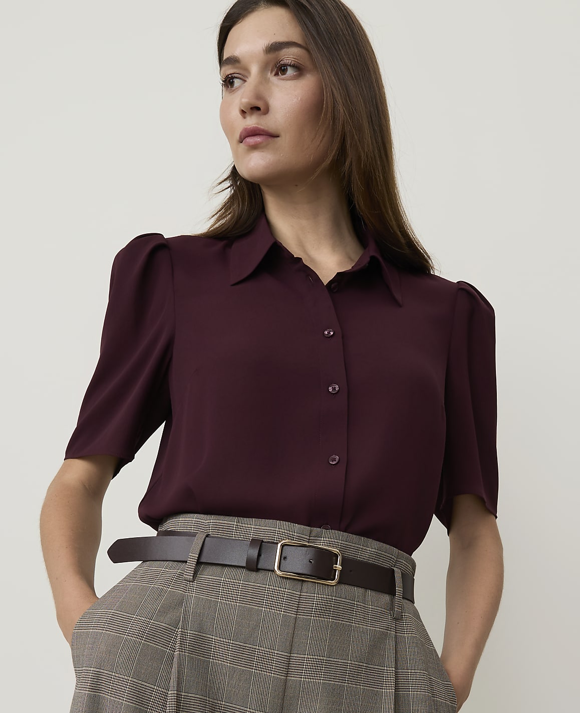 A woman in a burgundy blouse and plaid pants stands against a plain background.