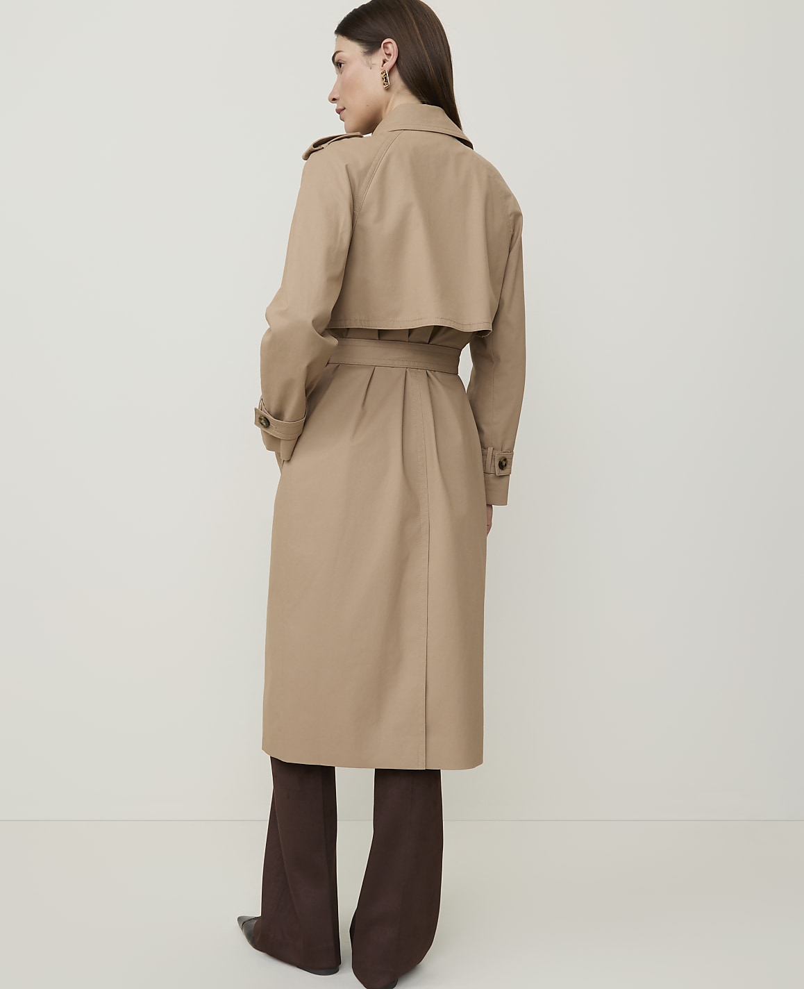 A woman in a tan trench coat and brown pants stands against a plain, light background.