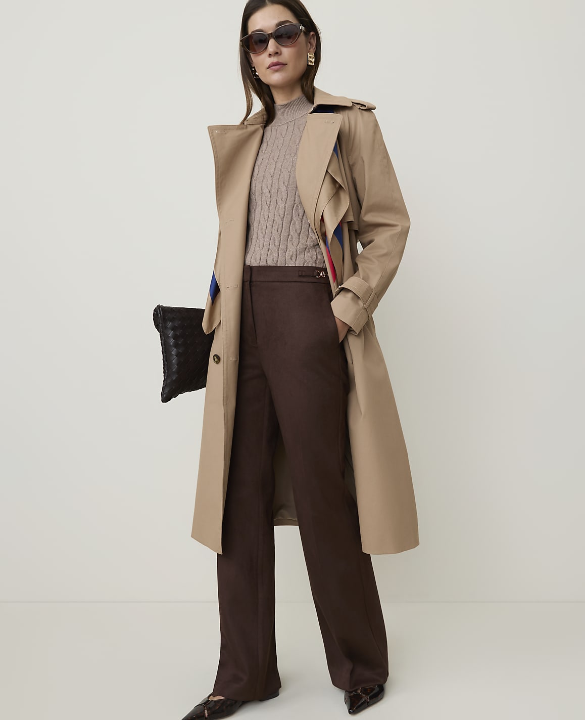 A woman in a tan trench coat and brown pants stands against a plain, light-colored background, holding a black clutch.