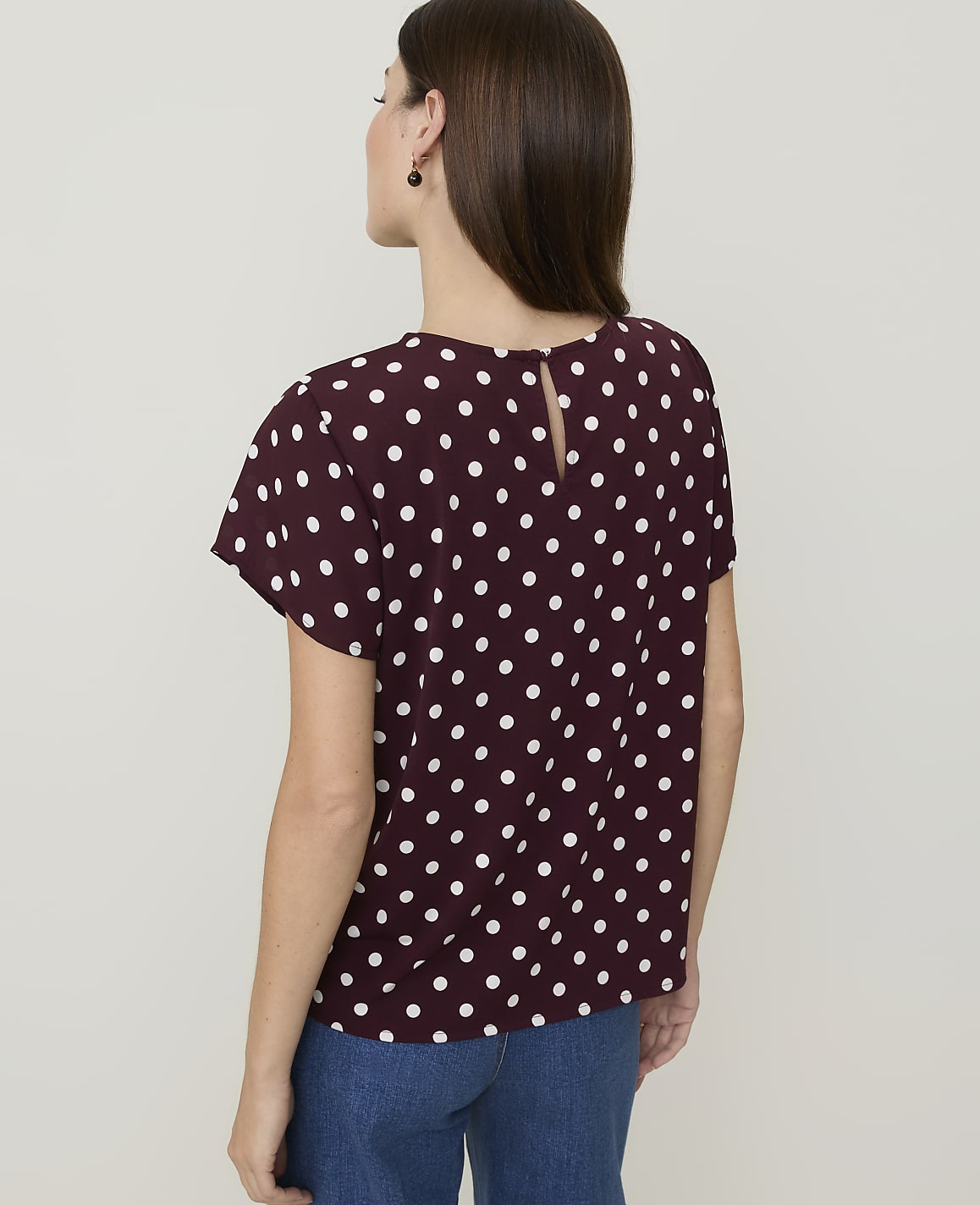 A woman in a burgundy polka dot top and blue jeans stands against a plain white background.