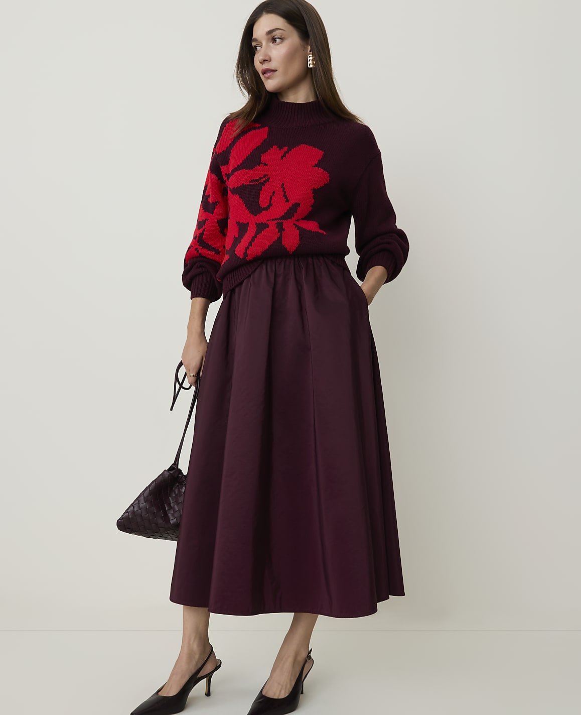 A woman in a burgundy sweater with a red floral print and a matching midi skirt stands against a plain, light-colored background, holding a dark handbag.