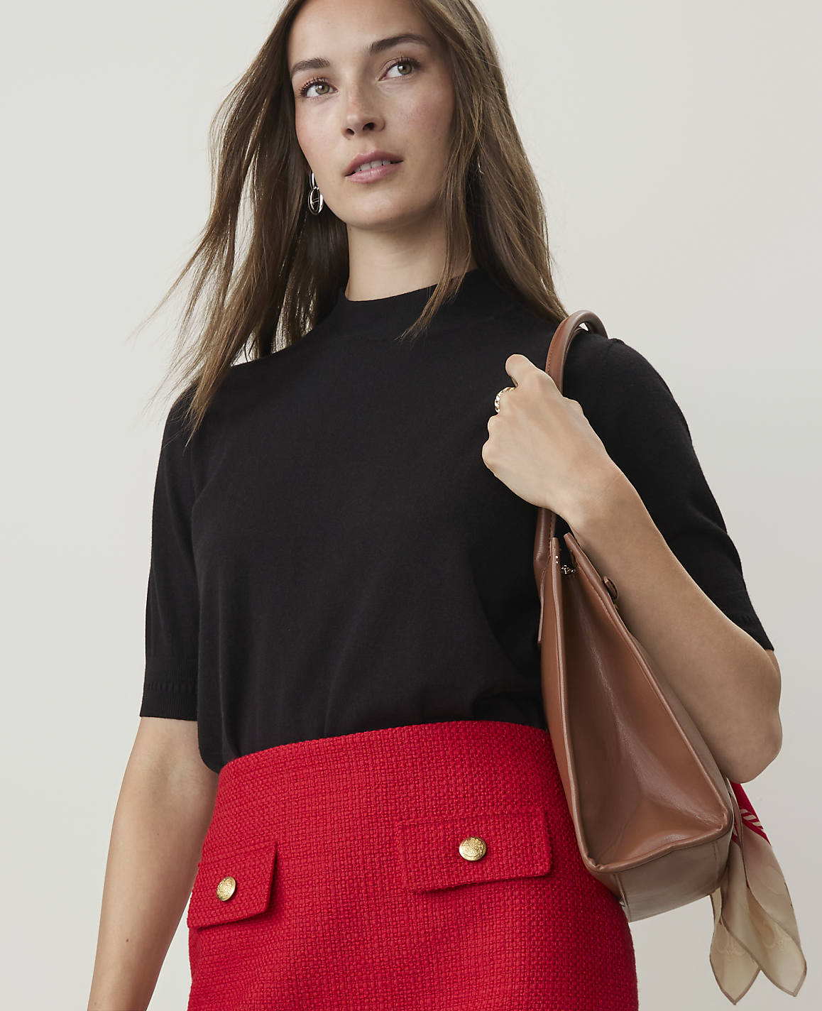 A woman in a black top and red skirt carries a brown purse against a plain background.