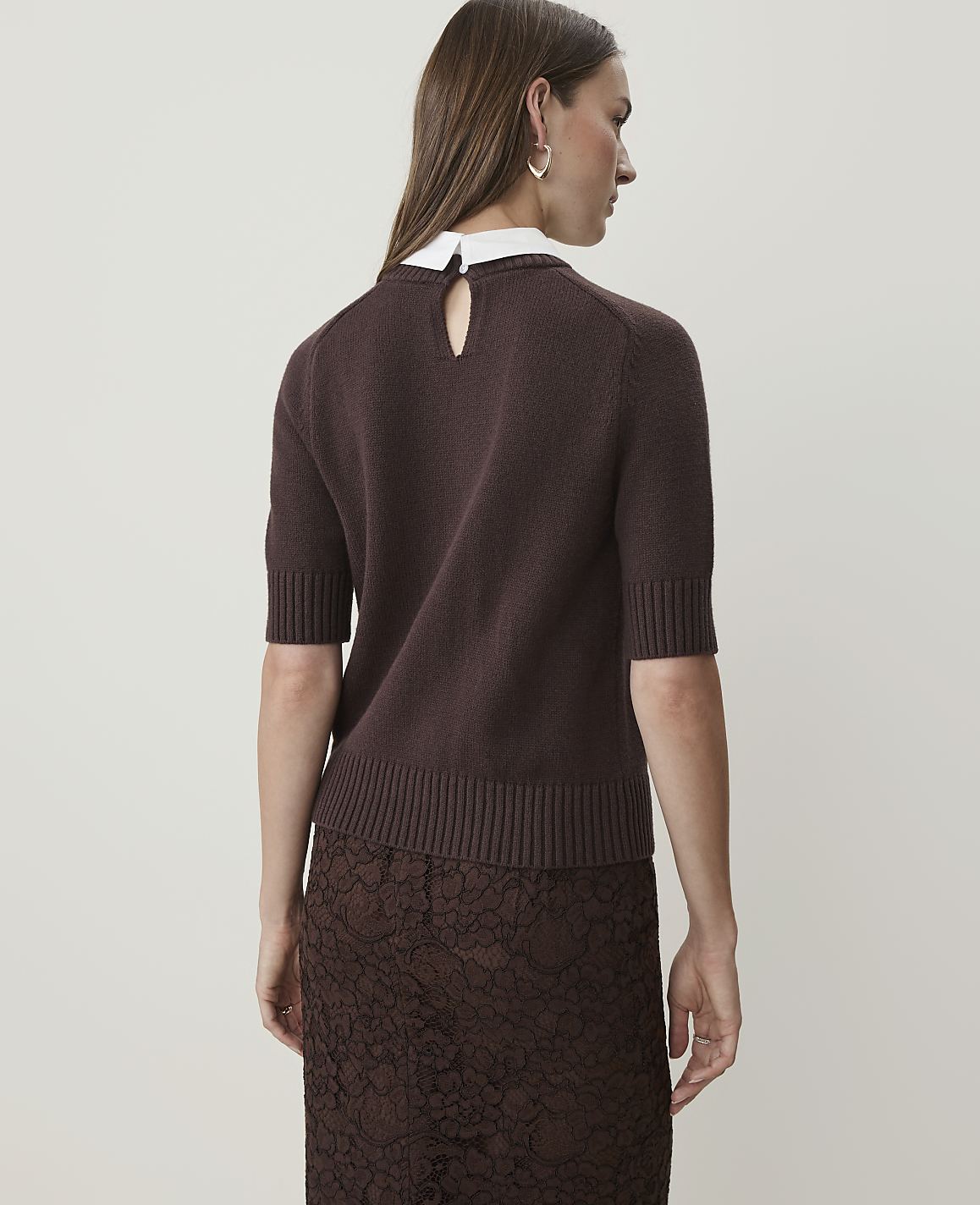A woman in a brown sweater and lace skirt stands against a plain white background.