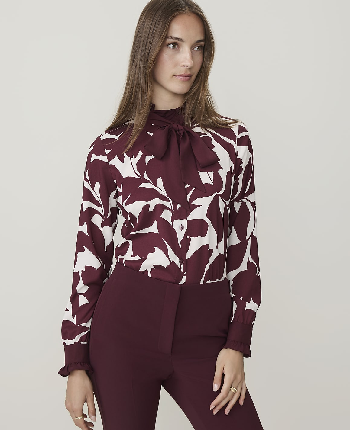 A woman in a maroon and white floral blouse and maroon pants stands against a plain, light gray background.