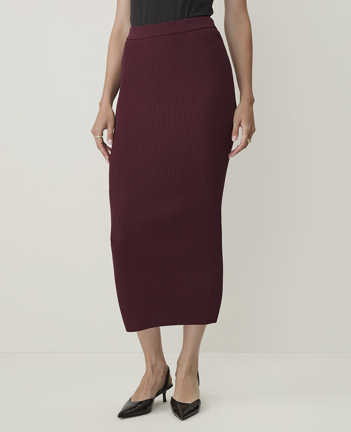 A person stands in a ribbed burgundy midi skirt and black slingback heels against a plain, light background.