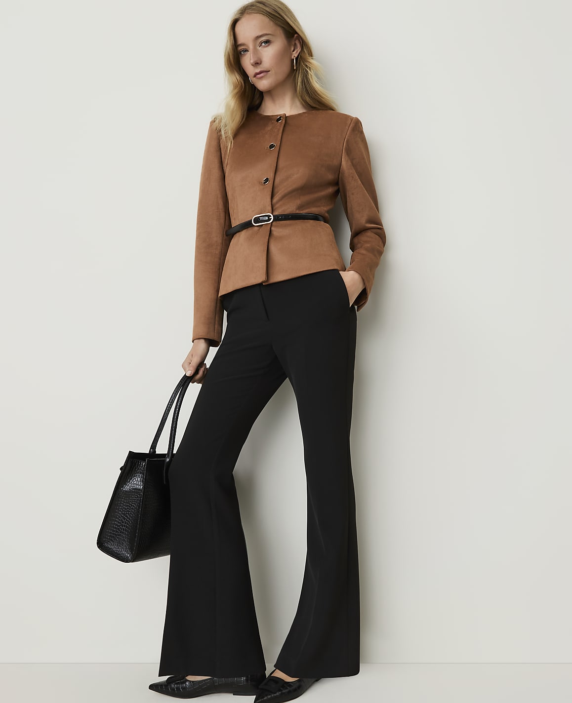 A woman in a brown jacket and black flared pants stands with a black handbag against a plain white background.