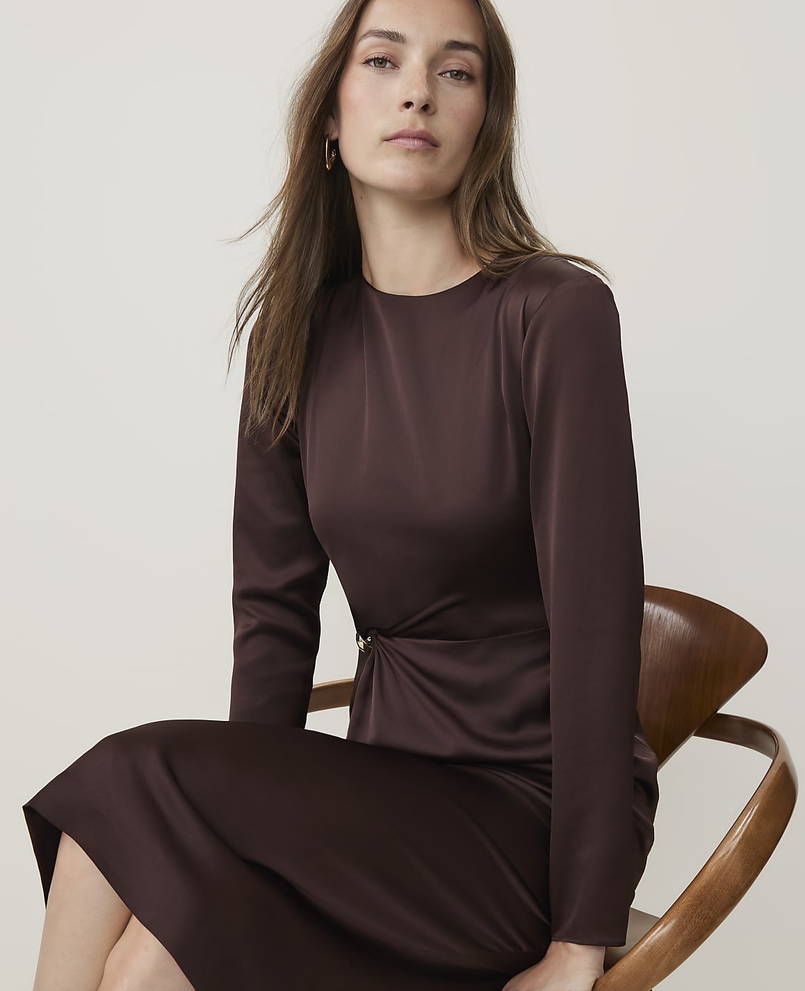 A woman in a brown satin dress with a twisted detail sits on a wooden chair against a plain white background.