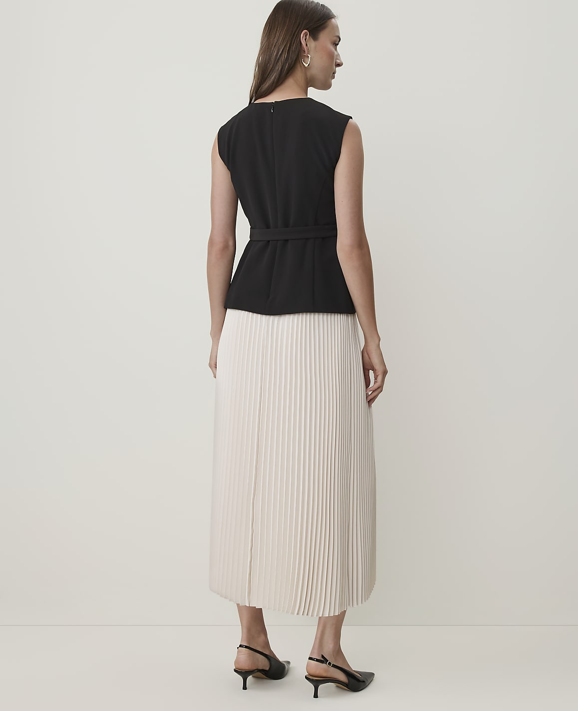 A woman in a black top and a pleated cream skirt stands against a plain white background.