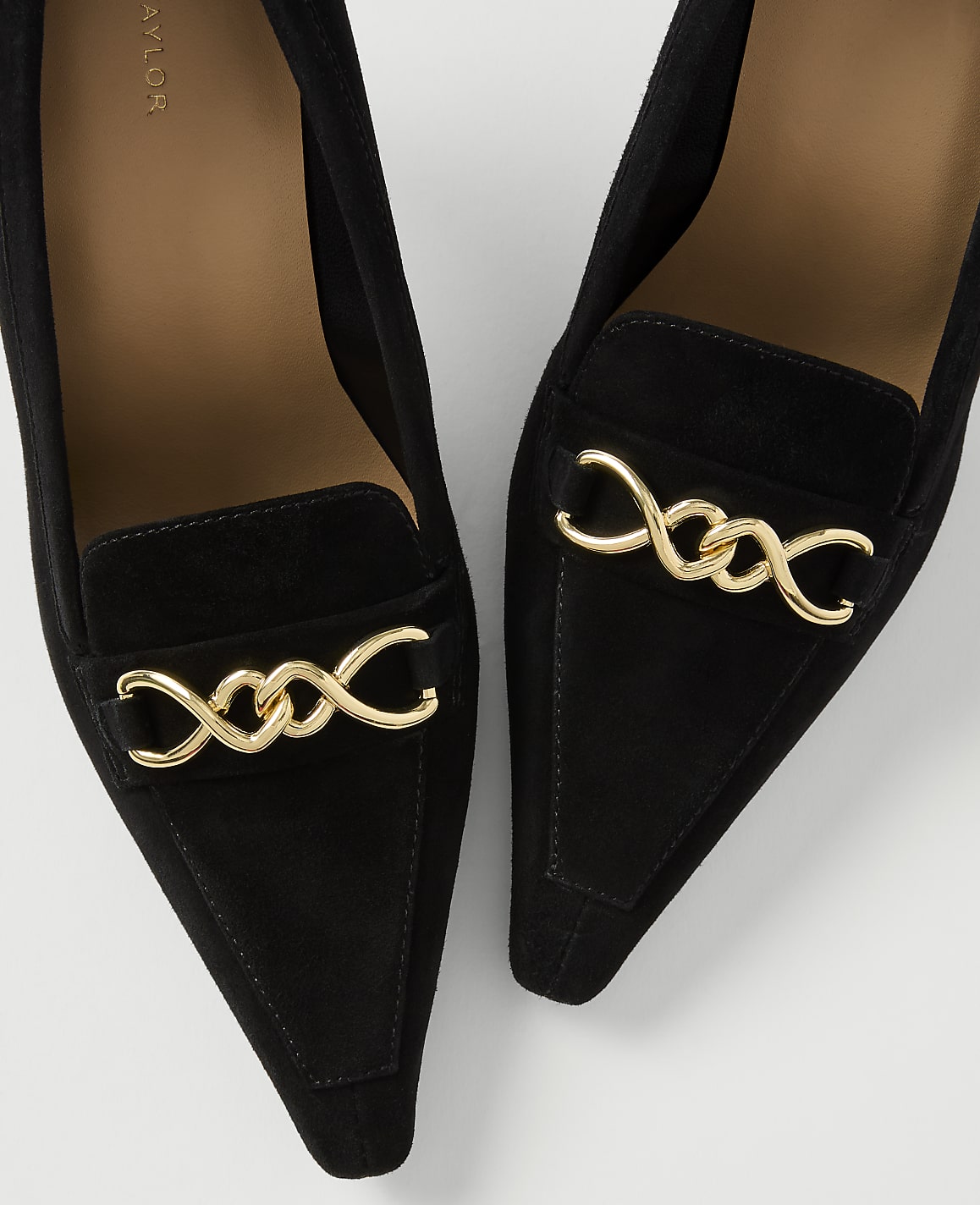A pair of black suede pointed-toe heels with gold chain accents are displayed against a plain white background.