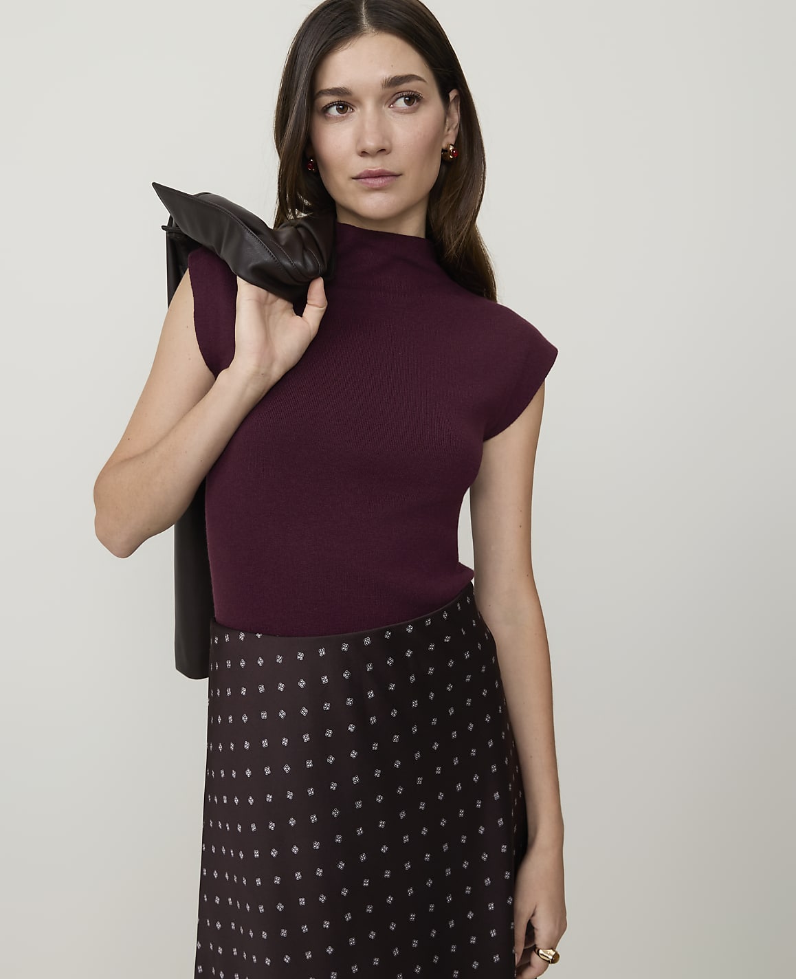 A woman in a burgundy mock-neck top and patterned skirt stands against a plain gray background, with a dark jacket draped over her shoulder.