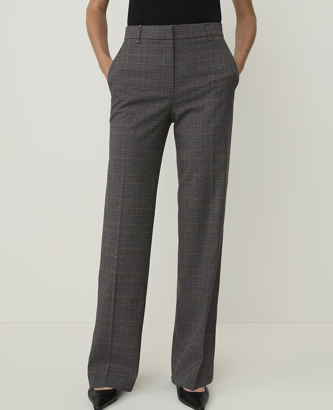 A person stands with hands in the pockets of their grey plaid wide-leg trousers against a plain white background.