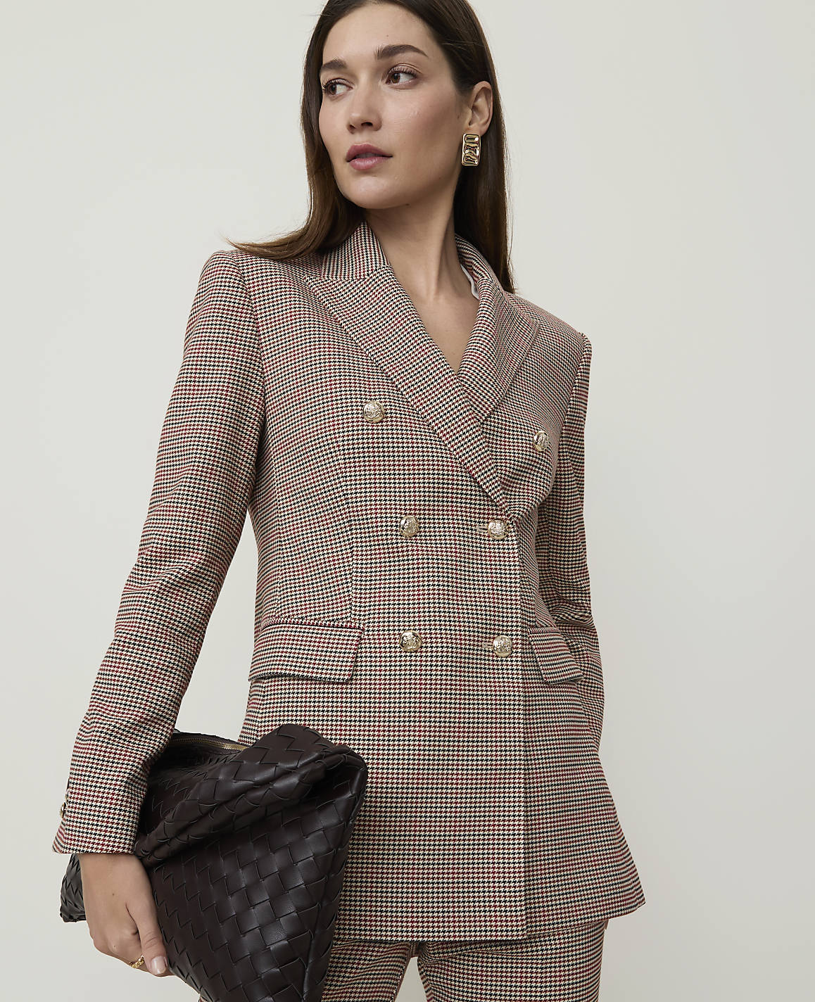 A woman in a patterned blazer and pants holds a woven clutch in front of a plain background.