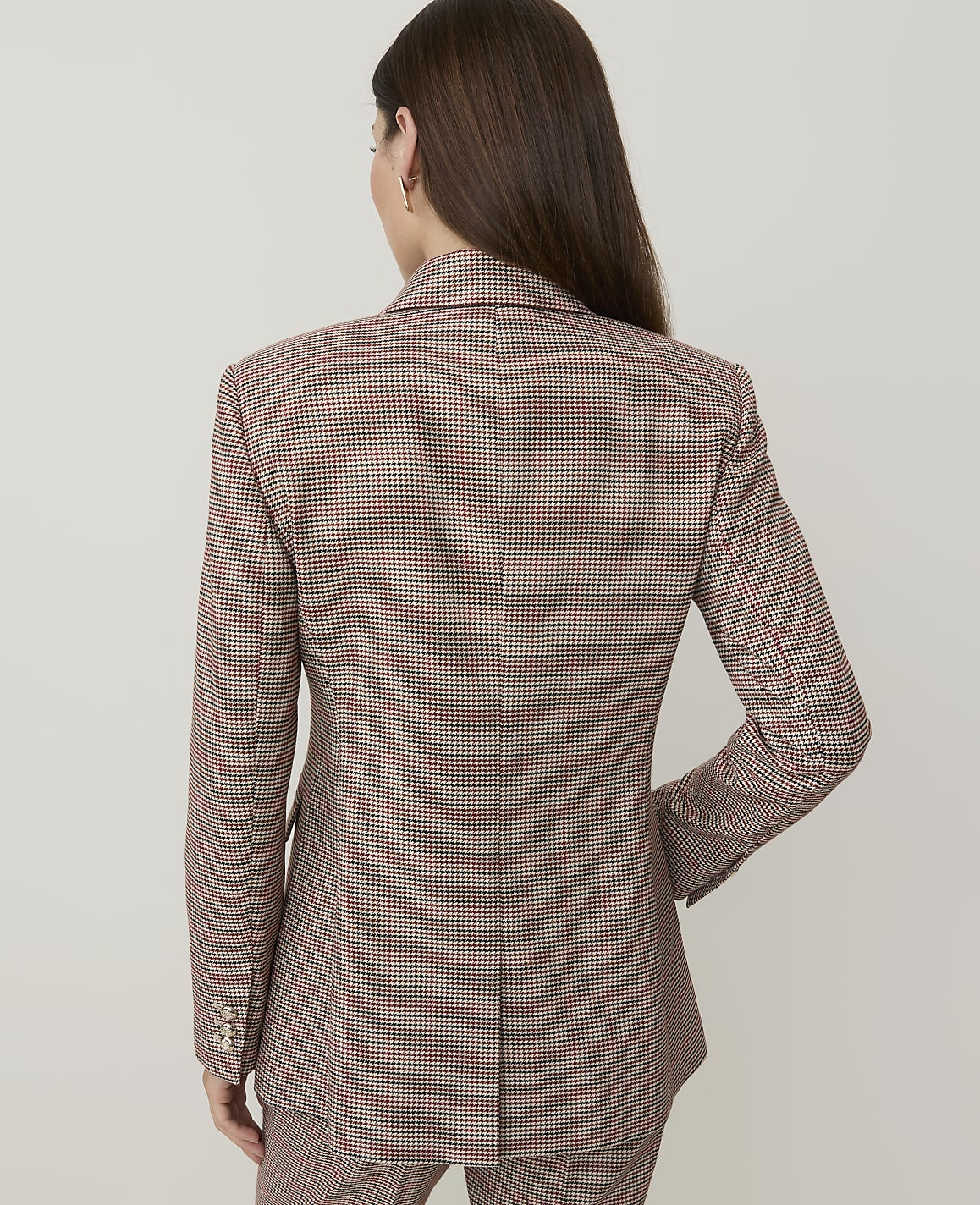 A woman in a brown and red checkered blazer stands against a plain white background.