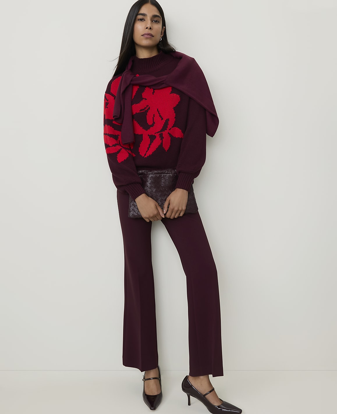 A woman in a burgundy sweater with a red floral print and matching pants holds a clutch in front of a plain white background.