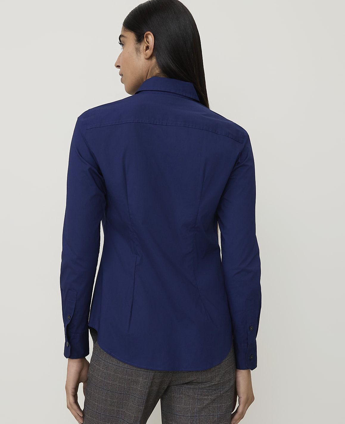 A woman in a dark blue collared shirt and plaid pants stands against a plain white background.