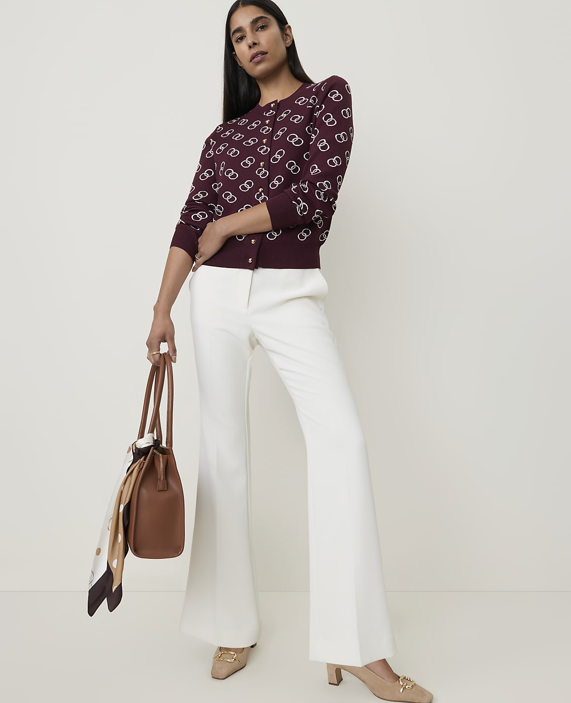 A woman in a patterned burgundy cardigan and white flared pants stands holding a brown handbag against a plain white background.